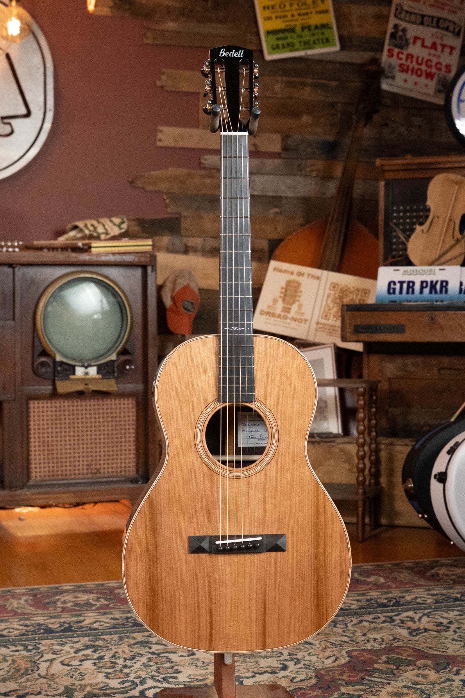 The Bedell Angelica Bellissima Red Cedar/Rosewood Parlor Guitar w/K&K Pure Mini Pickup #5002 by Bedell Guitars is shown upright in a cozy, music-filled room. Perfect for fingerstyle players seeking vintage charm and rich sound.