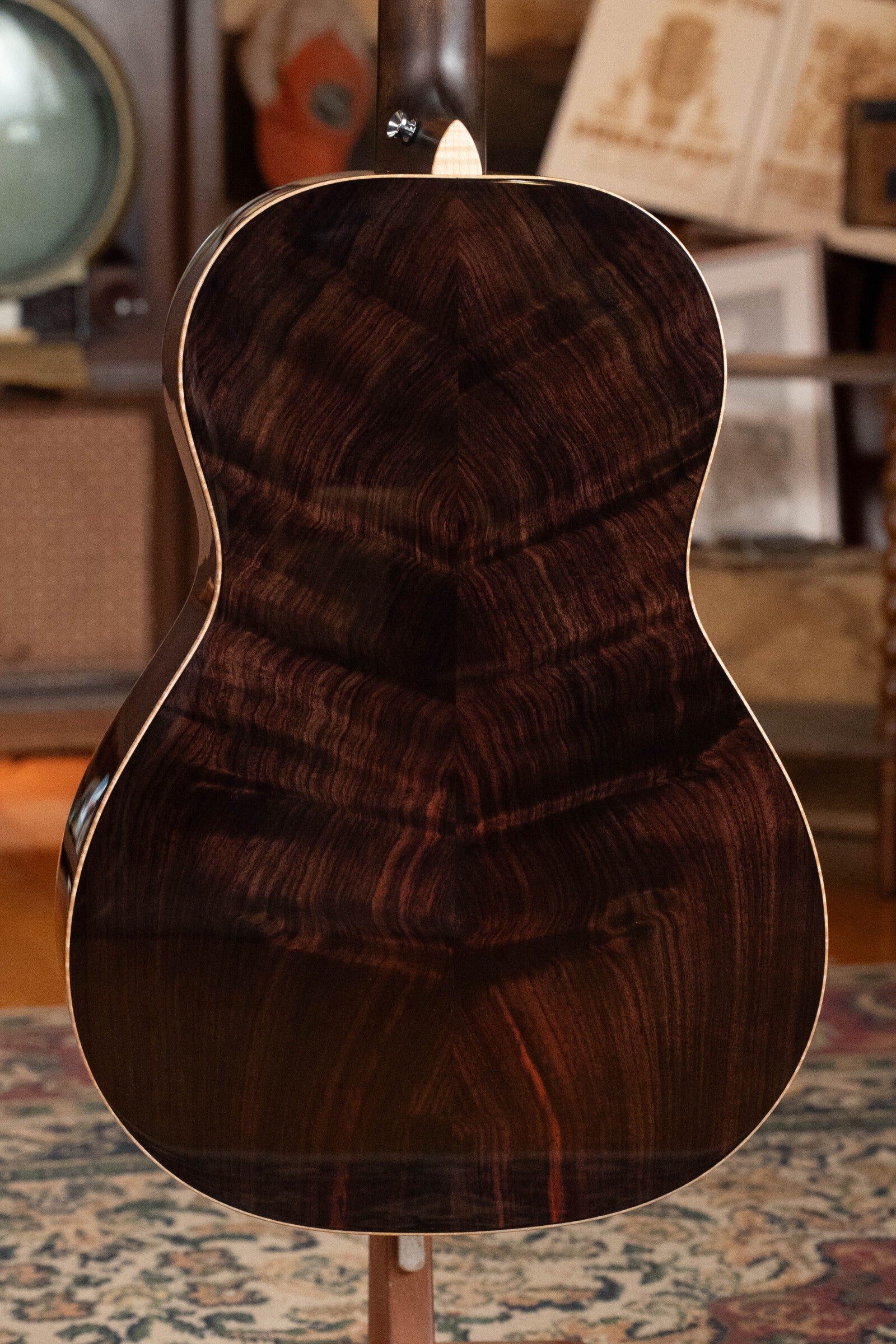 The image displays the back of a Bedell Angelica Bellissima Red Cedar/Rosewood Parlor Guitar w/K&K Pure Mini Pickup (#5002) by Bedell Guitars, showcasing its dark, polished wood and rich grain patterns on a patterned rug in a warmly lit room.