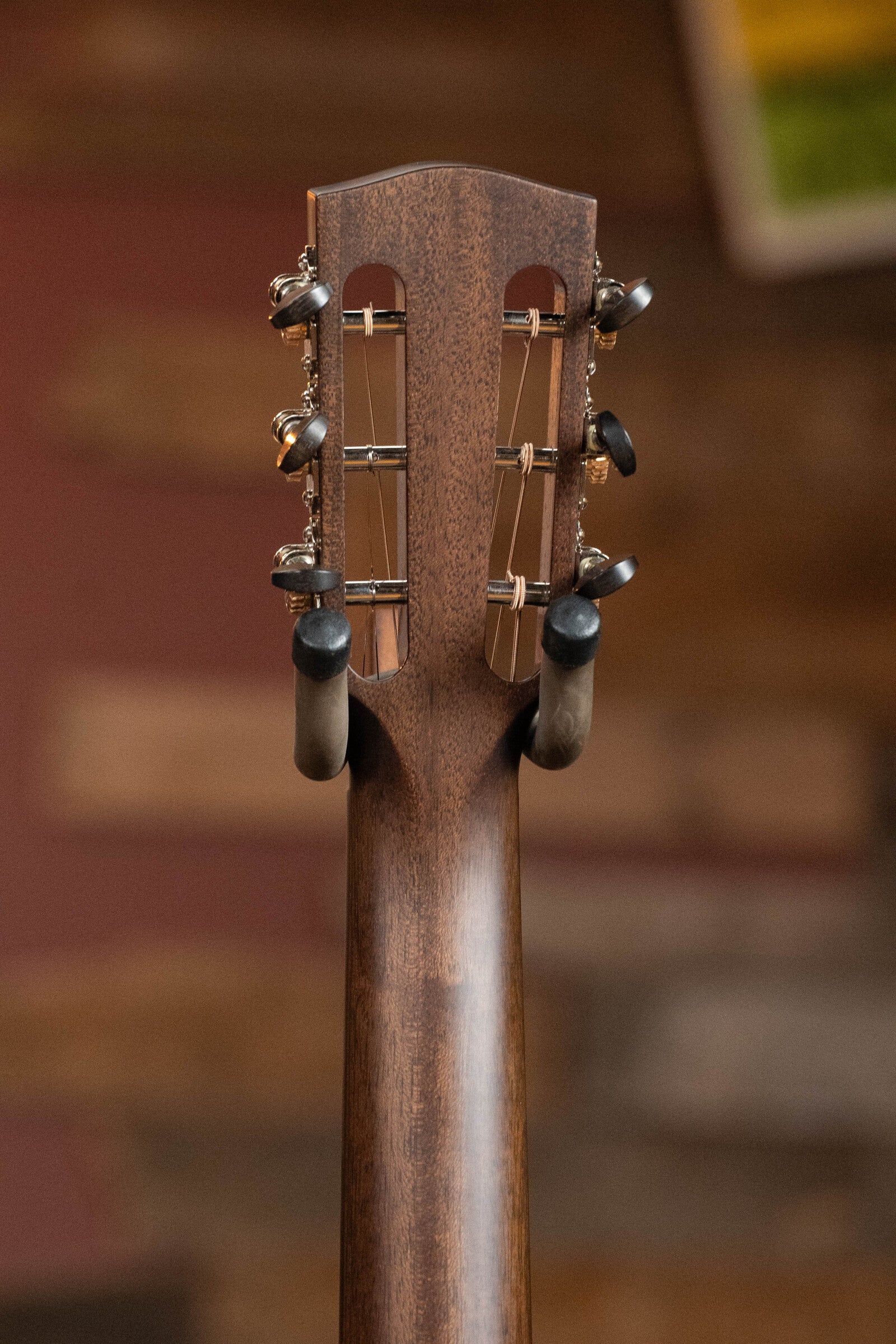 Close-up of the Bedell Guitars Angelica Bellissima Red Cedar/Rosewood Parlor Guitar w/K&K Pure Mini Pickup #5002 headstock, featuring metal tuners and held by a wall hanger against a blurred brown backdrop. Ideal for fingerstyle players.