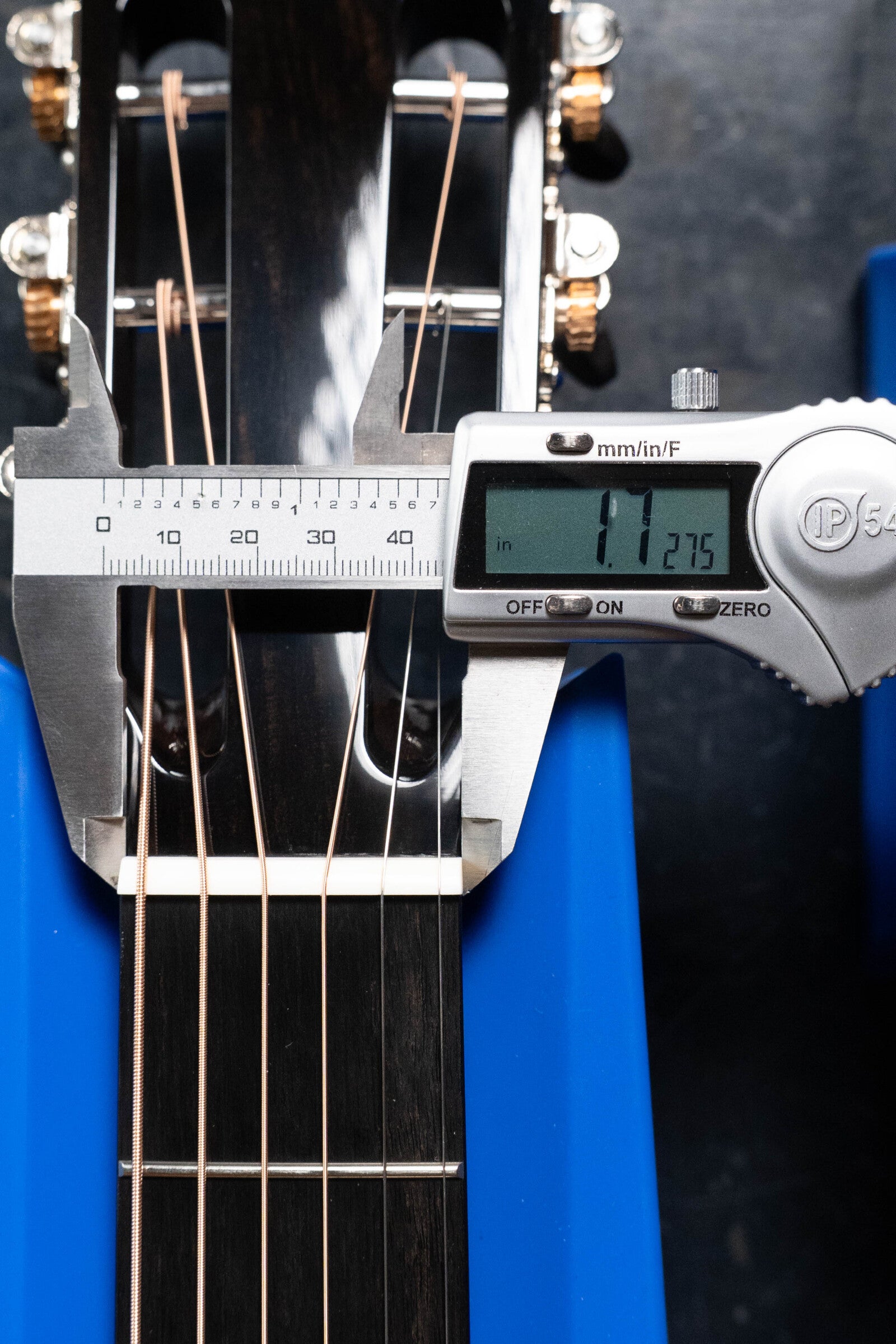 A digital caliper shows the Bedell Angelica Bellissima Red Cedar/Rosewood Parlor Guitar neck at the nut measures 1.275 inches. The Bedell Guitars instrument rests in a blue support cradle.