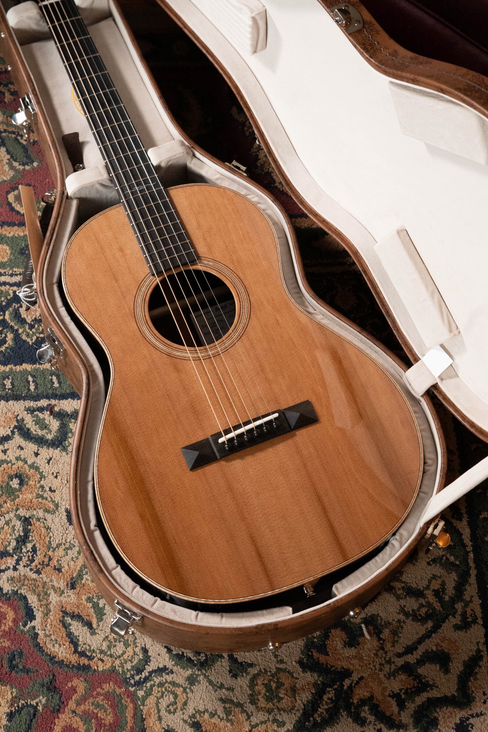 The Bedell Angelica Bellissima Red Cedar/Rosewood Parlor Guitar w/K&K Pure Mini Pickup (#5002) by Bedell Guitars rests in an open padded hard case on a patterned rug, its strings and fretboard perfect for fingerstyle players.