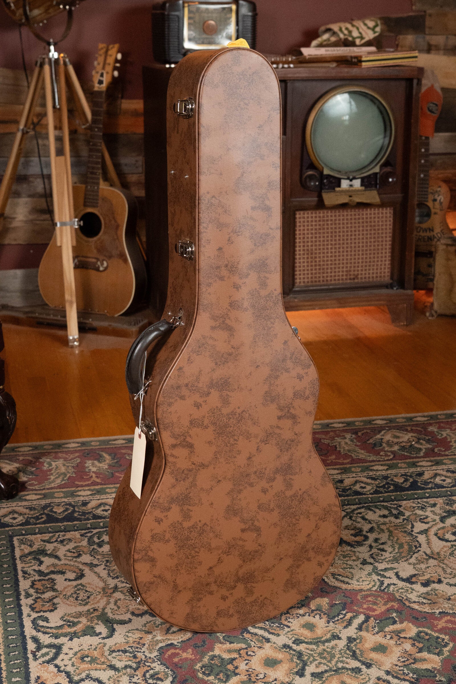 A Bedell Angelica Bellissima Red Cedar/Rosewood Parlor Guitar by Bedell Guitars stands upright in a cozy room with a patterned rug, vintage TV, tripod, and fingerstyle guitar in the background.