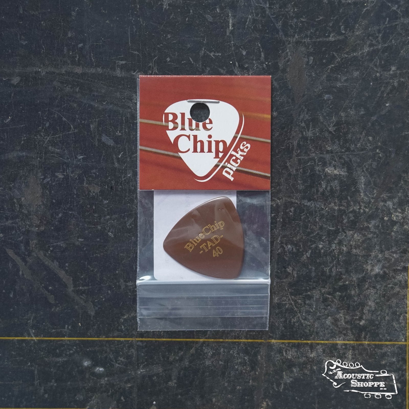 A packaged BlueChip Picks (TAD40) guitar pick rests on a dark, textured surface. The packaging shows the BlueChip brand logo in red and white and includes The Acoustic Shoppe logo, ideal for acoustic accessory collections.