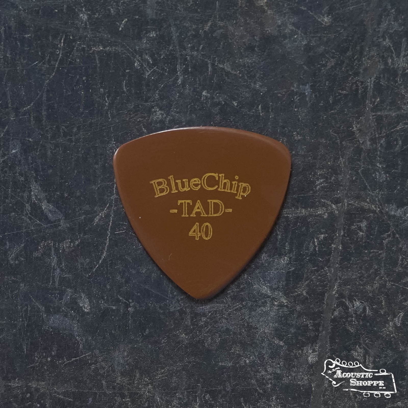 A BlueChip Picks (TAD40) guitar pick by BlueChip, featuring gold lettering and a brown finish, rests on a dark marbled surface, highlighting high-quality acoustic accessories.