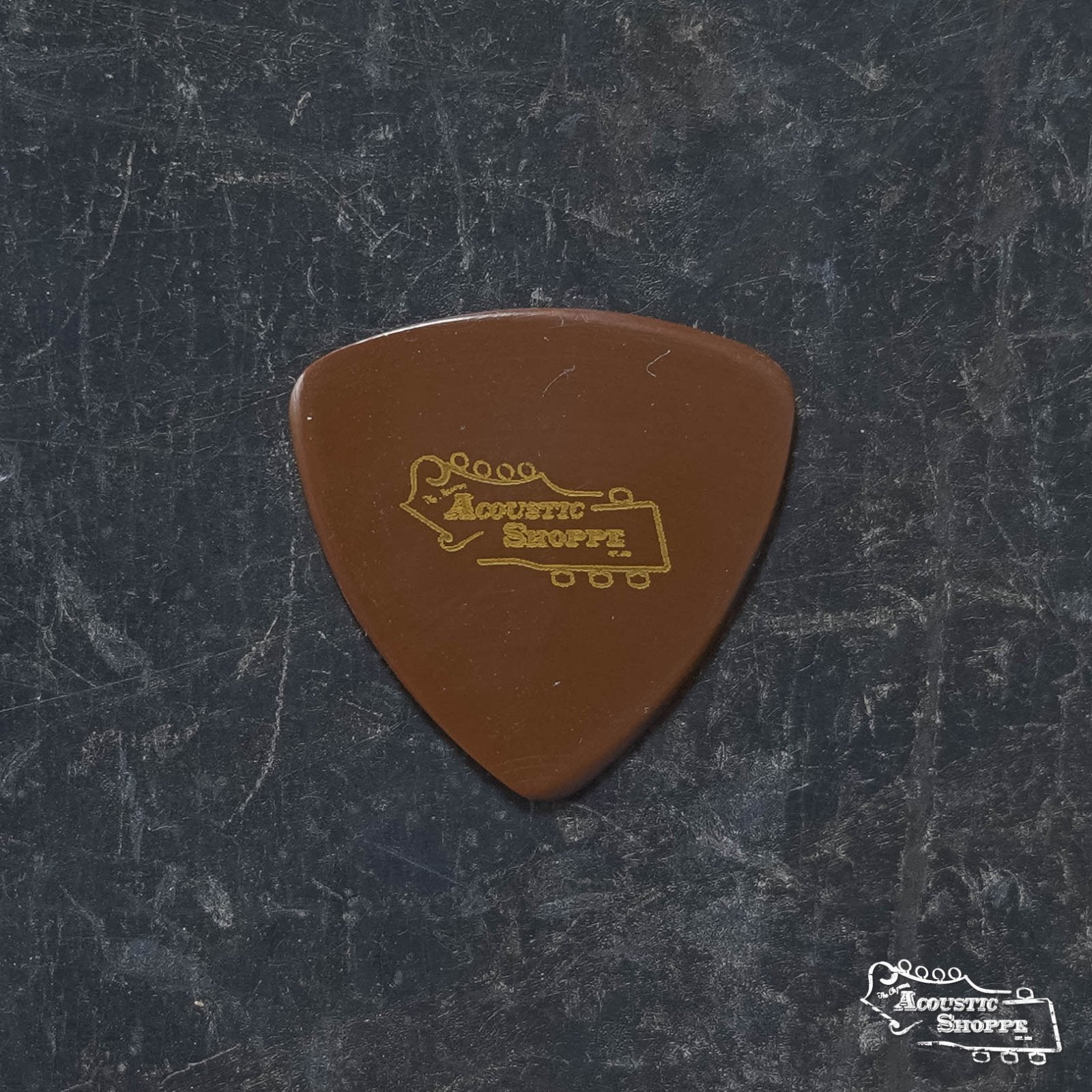 A BlueChip Picks (TAD40) guitar pick from BlueChip, featuring the Acoustic Shoppe logo in gold, rests on a dark, textured surface. The high-quality pick stands out, with the same logo shown in white at the image's bottom right.