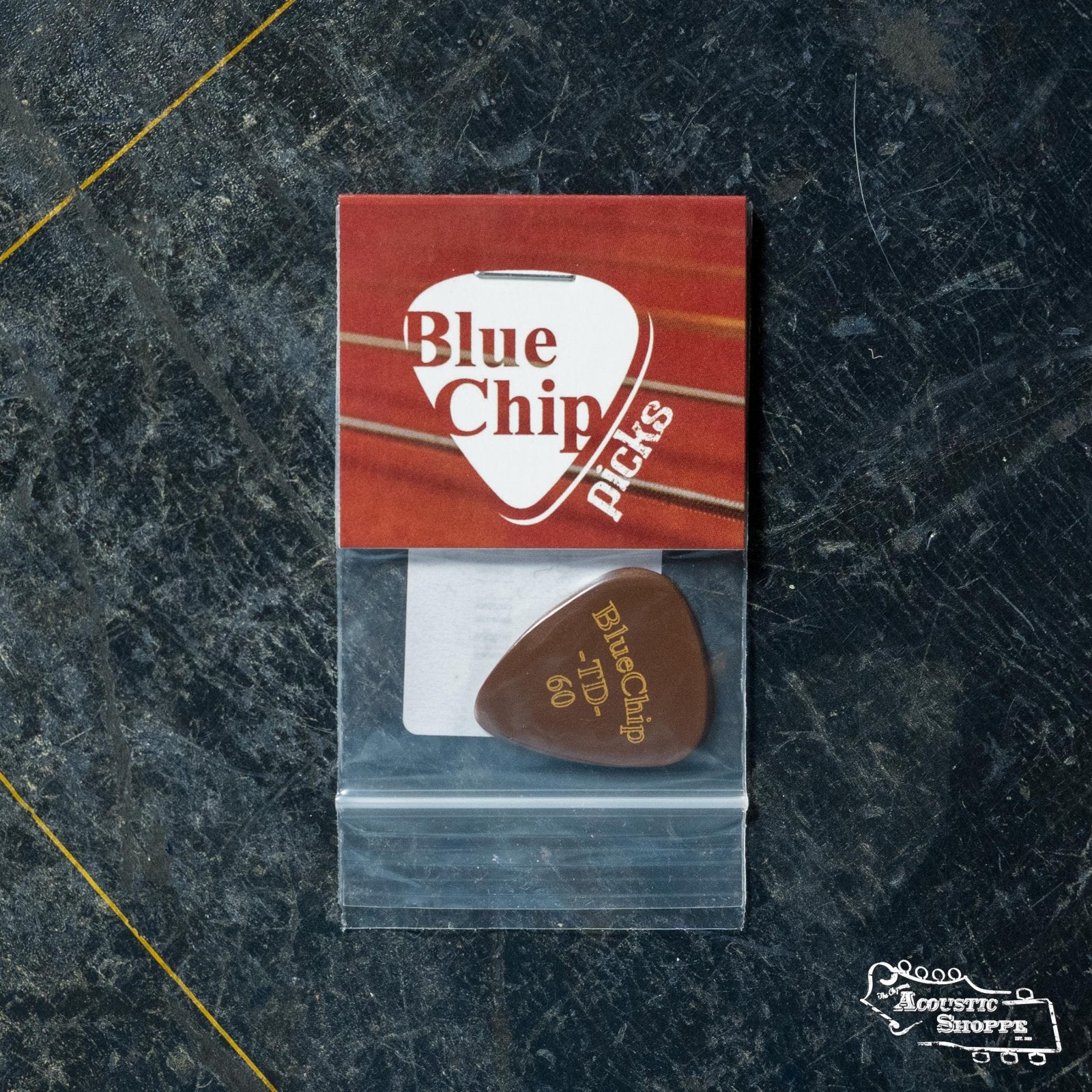 A BlueChip Picks (TD60) teardrop guitar pick by BlueChip, in a small plastic bag with a red and white label, sits on a dark textured surface with a yellow diagonal line and the Acoustic Shoppe logo in the corner.