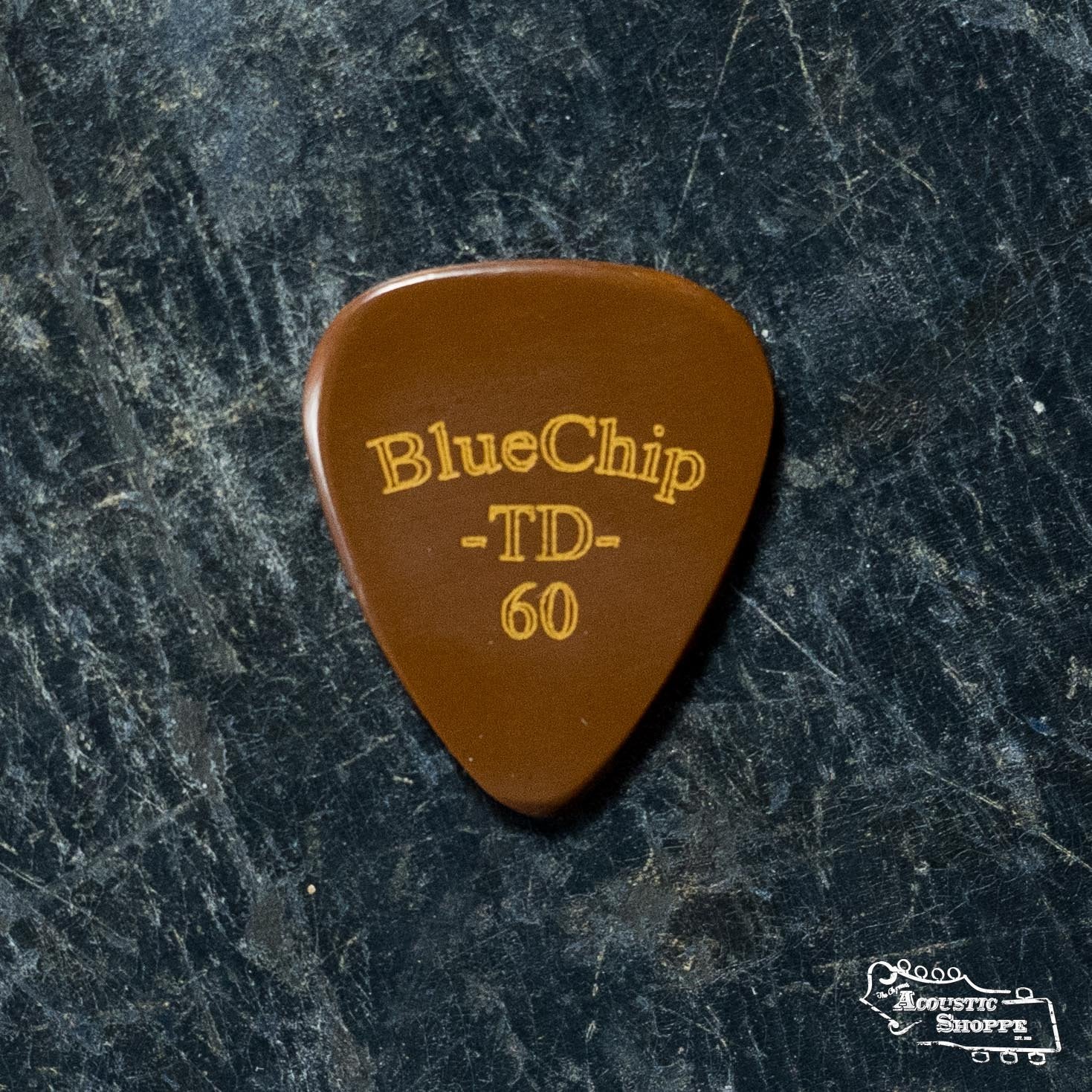A brown BlueChip Picks (TD60) pick with the brand name in gold rests on a dark, textured surface. The Acoustic Shoppe logo, representing an acoustic instrument dealer, appears in the lower right corner.