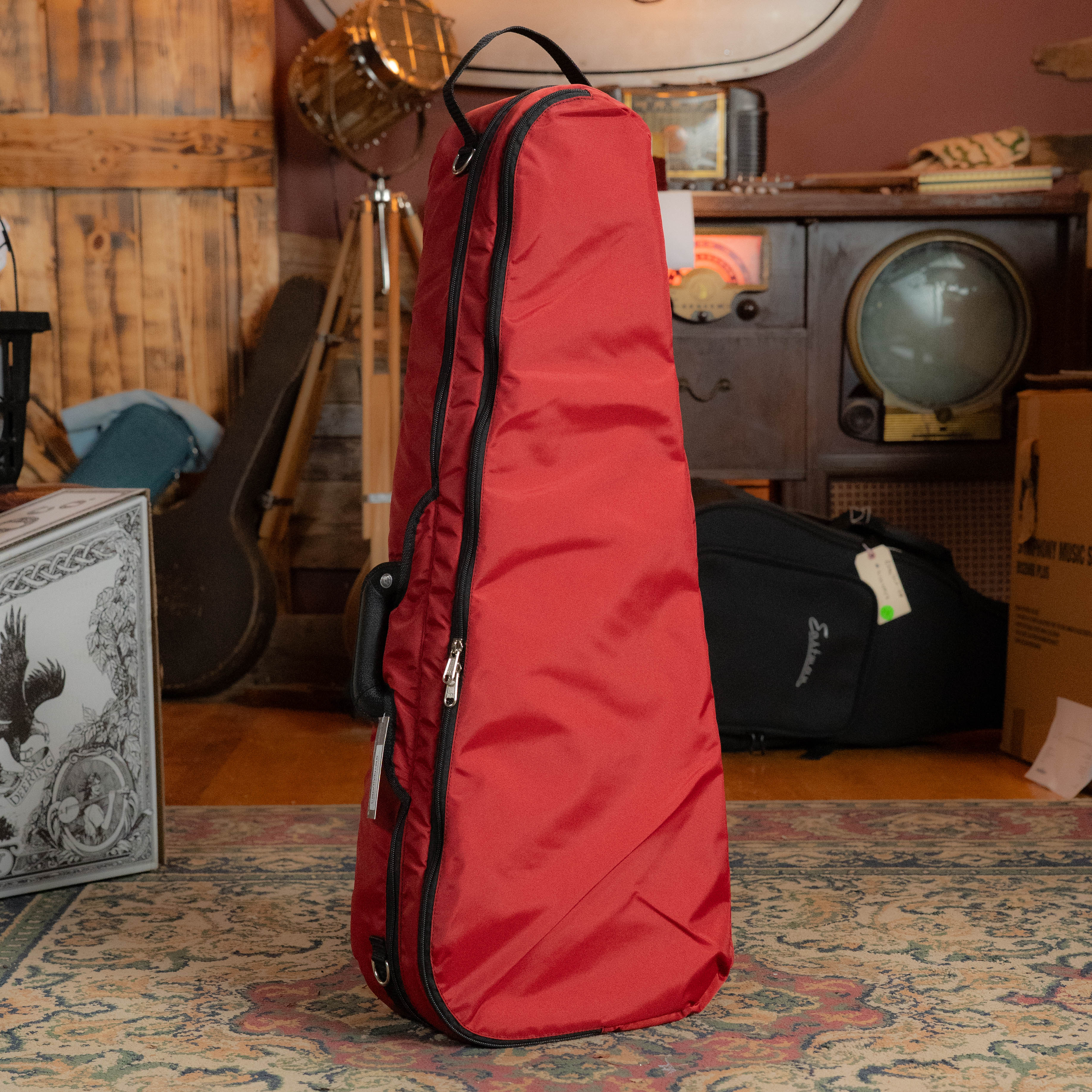 A Howard Core Bobelock Arrow Red/Gray F-Style Fiberglass Mandolin Case with a handle stands upright on a patterned rug in a cozy, vintage-inspired room filled with musical equipment.