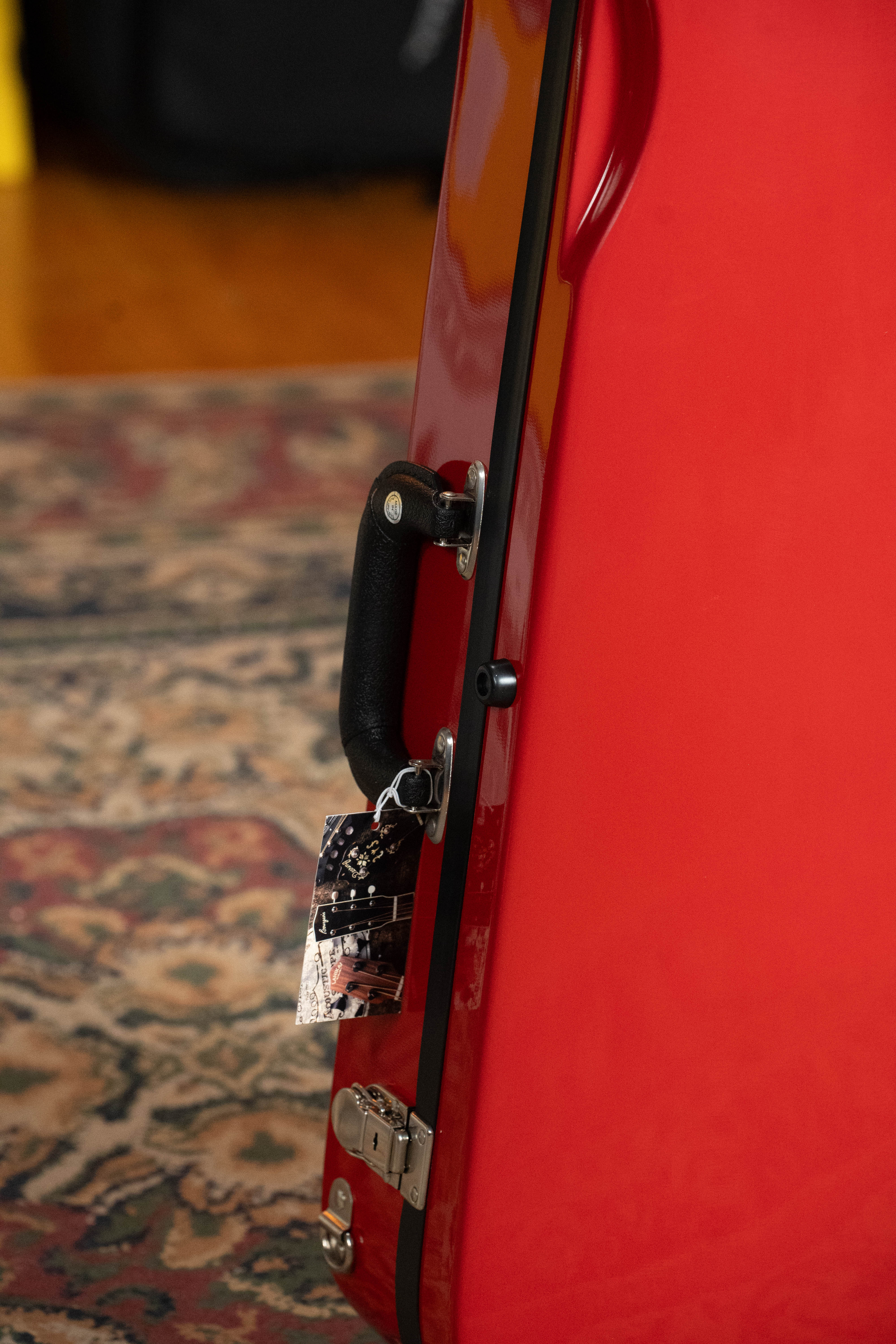 A close-up of the Howard Core Bobelock Arrow Red/Gray F-Style Fiberglass Mandolin Case stands upright on a patterned rug, highlighting its black handle and attached metal key tag.
