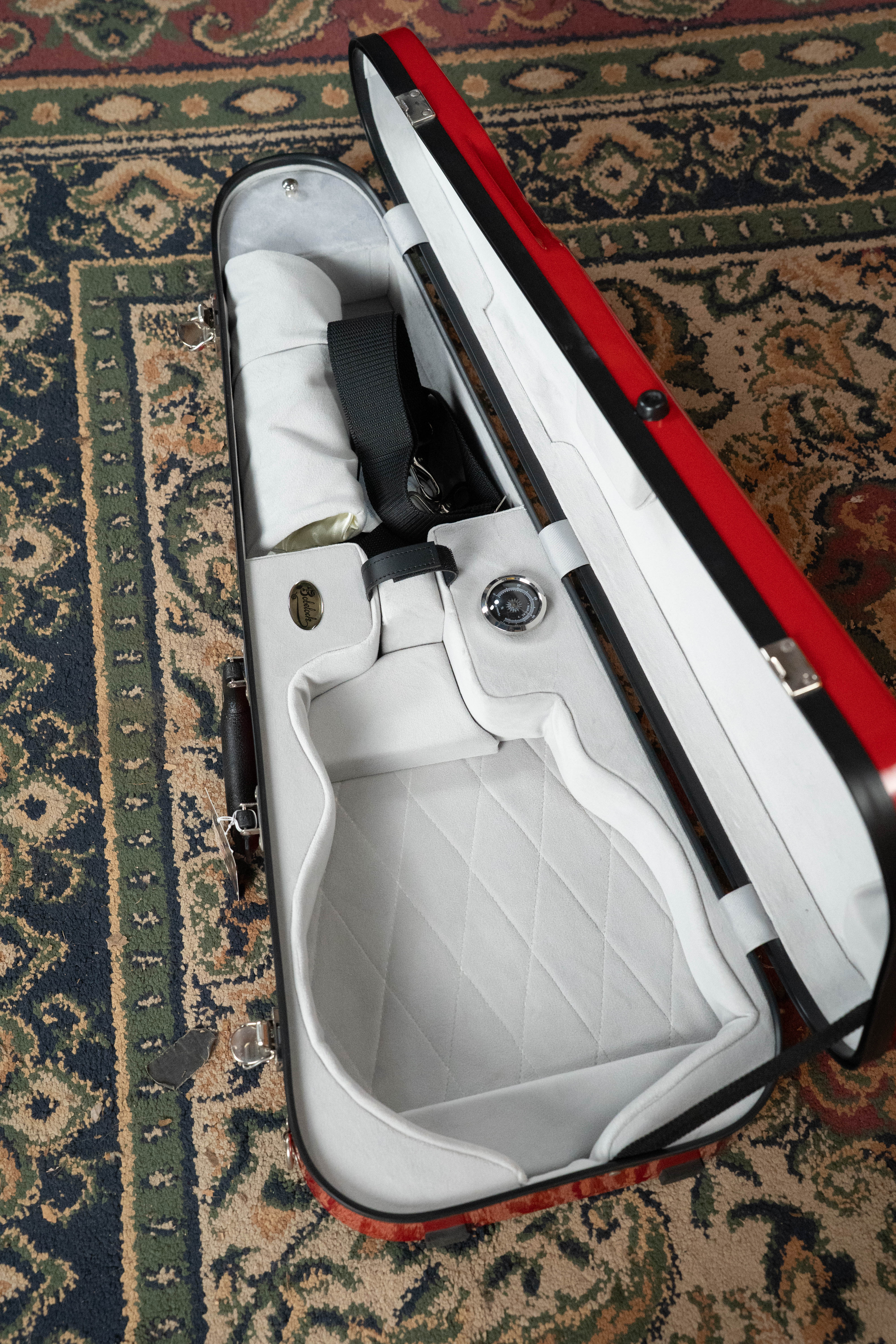 A Bobelock Arrow Red/Gray F-Style Fiberglass Mandolin Case by Howard Core, with a soft quilted white interior, sits open on a patterned rug. Its arrow-shaped exterior includes sections for a mandolin, bow, and accessories.