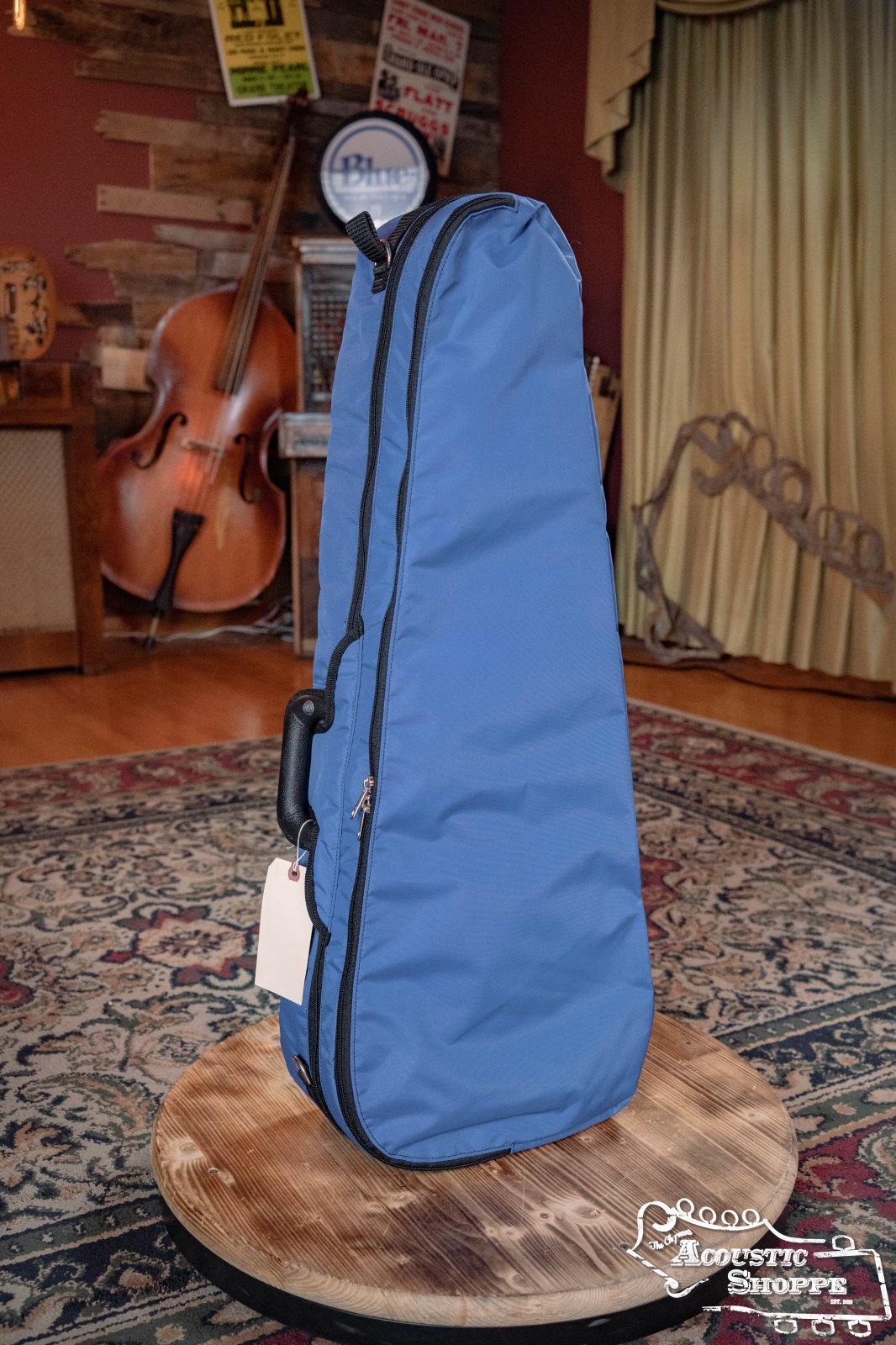 A Bobelock Arrow Blue/Gray Fiberglass F-Style Mandolin Case (B6-1029FBLS-BUGY) stands upright on a round wooden table in a cozy room. The case features a handle, zipper, and pouch for spare cable—connector not included.