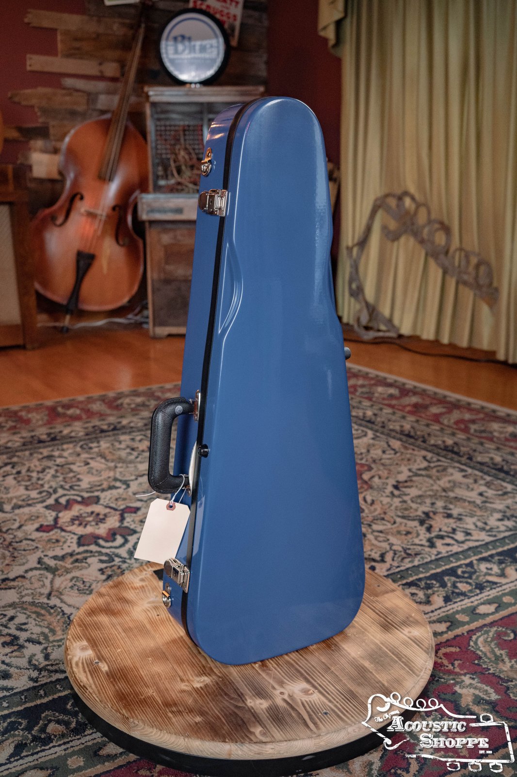 A Bobelock Arrow Blue/Gray Fiberglass F-Style Mandolin Case (B6-1029FBLS-BUGY) stands upright on a round wooden table with a double bass and rustic decor in the background. The Acoustic Shoppe logo appears in the lower right corner.