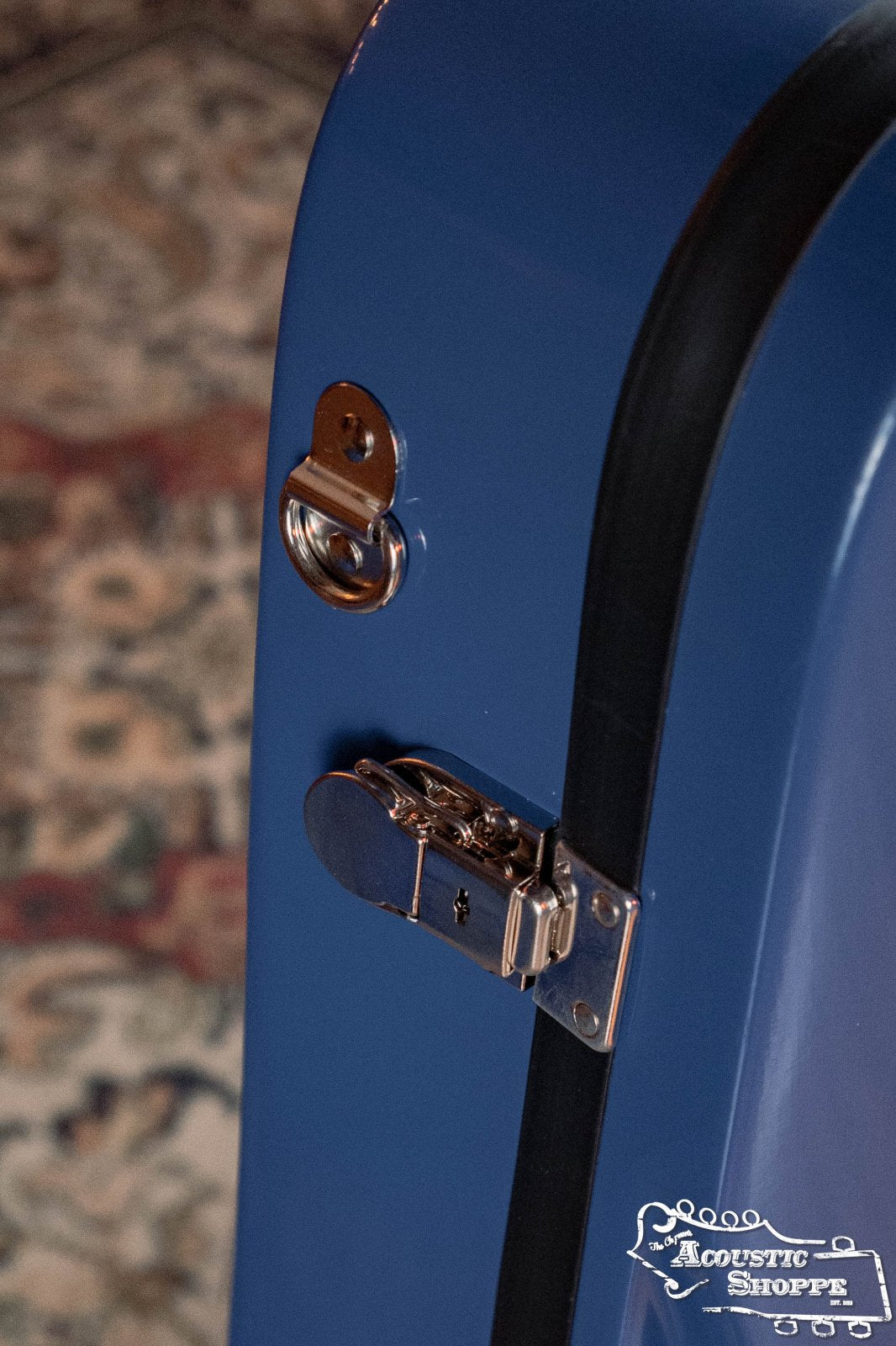 A close-up of the Bobelock Arrow Blue/Gray Fiberglass F-Style Mandolin Case (B6-1029FBLS-BUGY) with a metal latch and lock, placed on a patterned rug. The Acoustic Shoppe logo is visible; no connector or spare cable is shown.