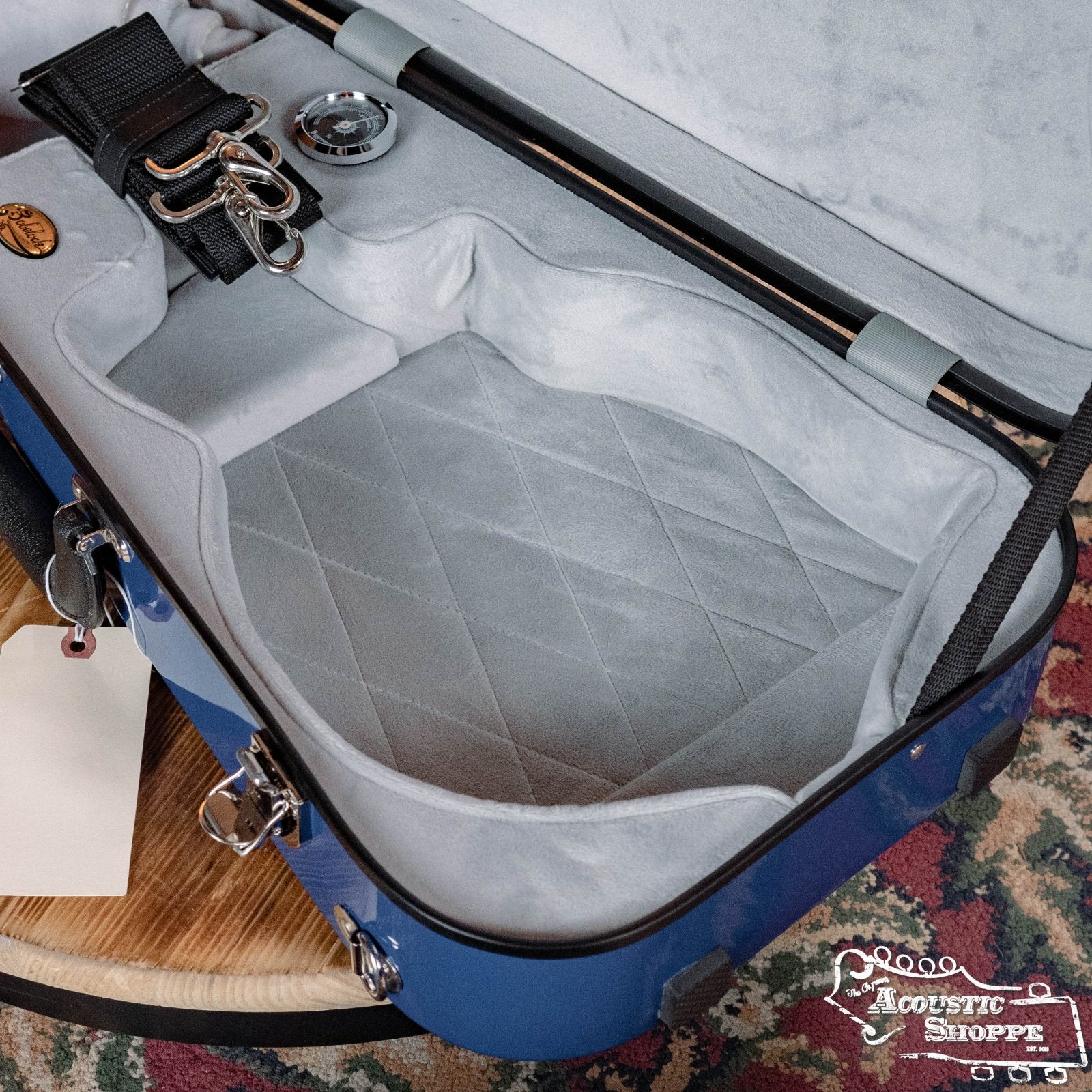 The Bobelock Arrow Blue/Gray Fiberglass F-Style Mandolin Case (B6-1029FBLS-BUGY) features a soft quilted gray interior, metal latches, and a shoulder strap. Includes a spare cable (no connector attached). The Acoustic Shoppe logo appears in the lower right corner.