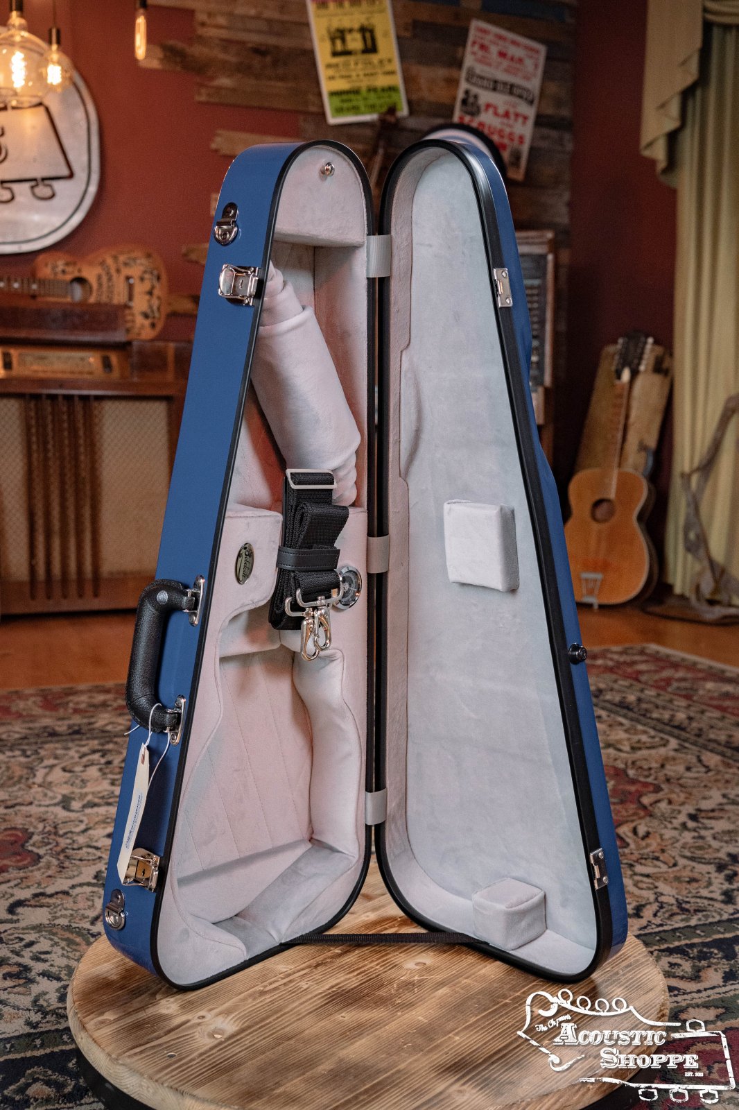 A blue and gray Bobelock Arrow Fiberglass F-Style Mandolin Case (B6-1029FBLS-BUGY) stands open on a patterned rug in a cozy, rustic room with several guitars and wooden furniture. The Acoustic Shoppe logo appears in the lower right corner.