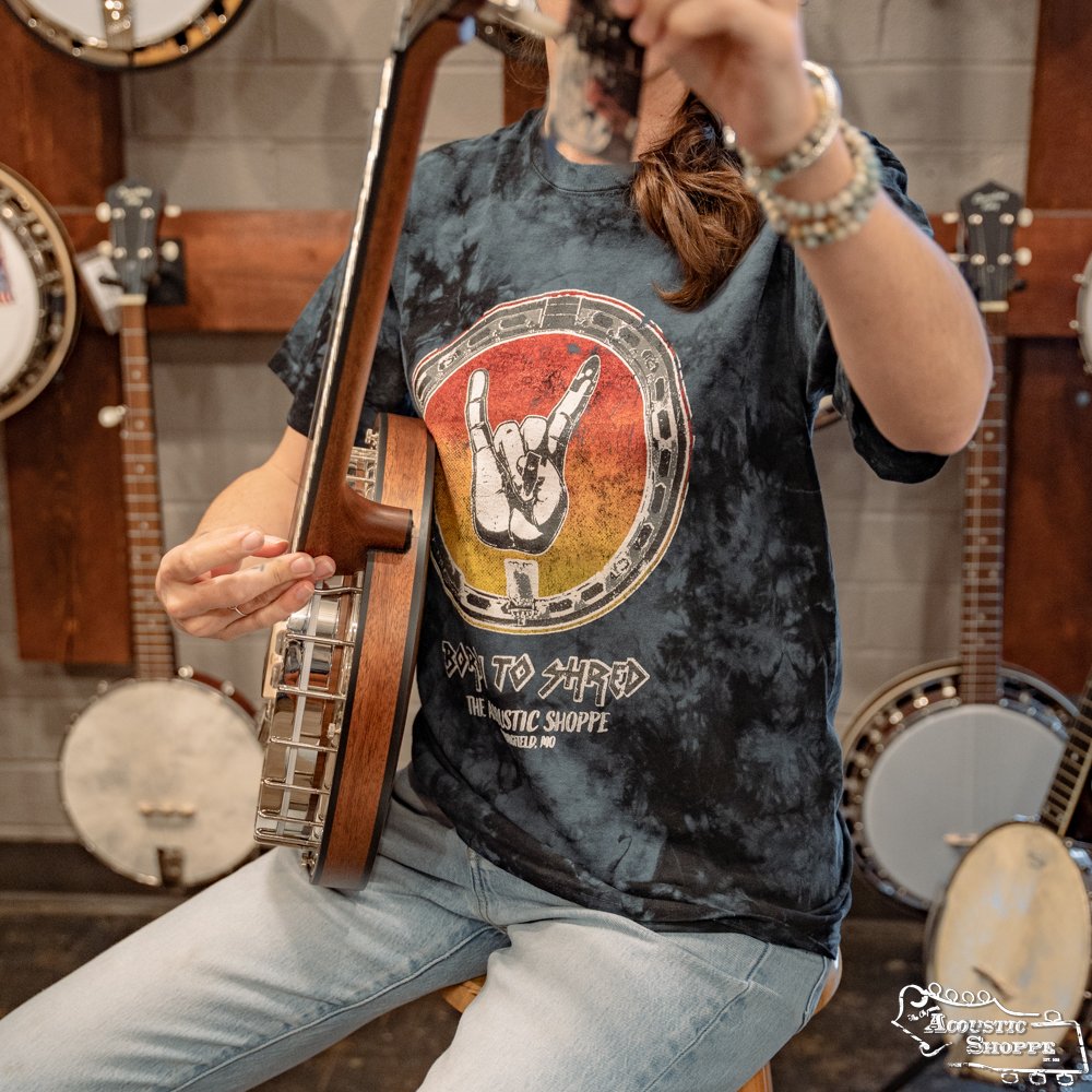 A person wearing an Amplify Screen Printing & Promotions “Banjo Shred” Design T-Shirt from The Acoustic Shoppe plays a banjo, with several other banjos like those at Acoustic Shoppe hanging in the background. The persons face isn’t visible.