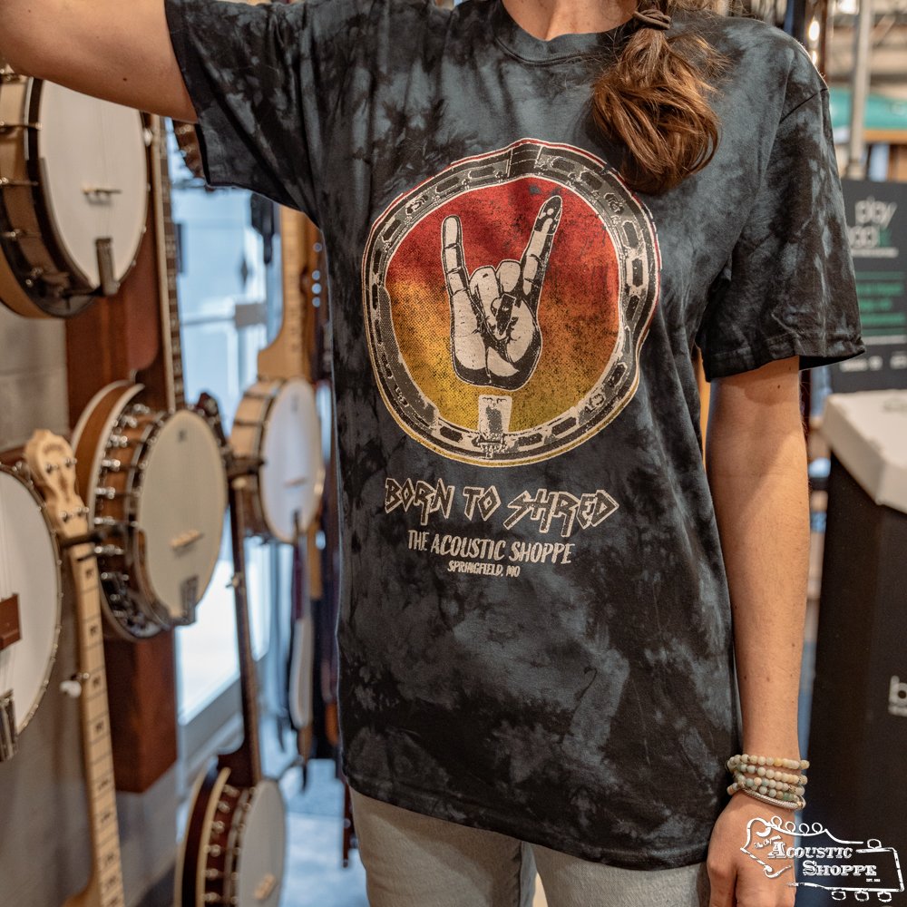Wearing The Acoustic Shoppe Banjo Shred Design T-Shirt by Amplify Screen Printing & Promotions, a person throws the rock on sign inside a round graphic reading Born to Shred, with several Eastman banjos visible in the background.