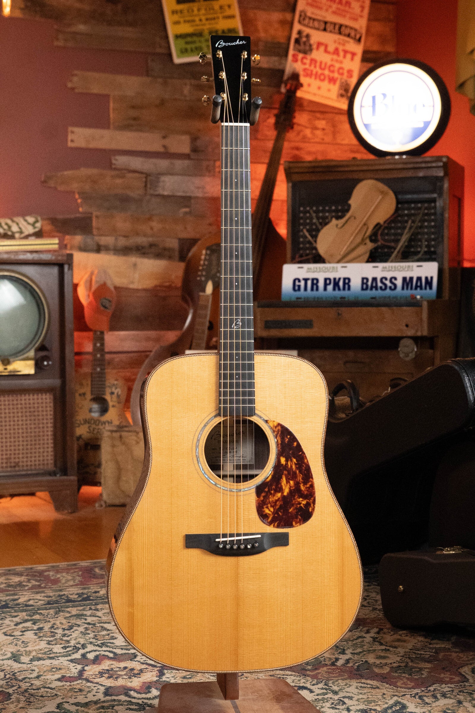 The Boucher BG-52-GM Bluegrass Goose Dreadnought with a torrefied Adirondack red spruce top stands on a rug in a cozy, vintage room with wooden walls, music gear, and retro signs in the background.