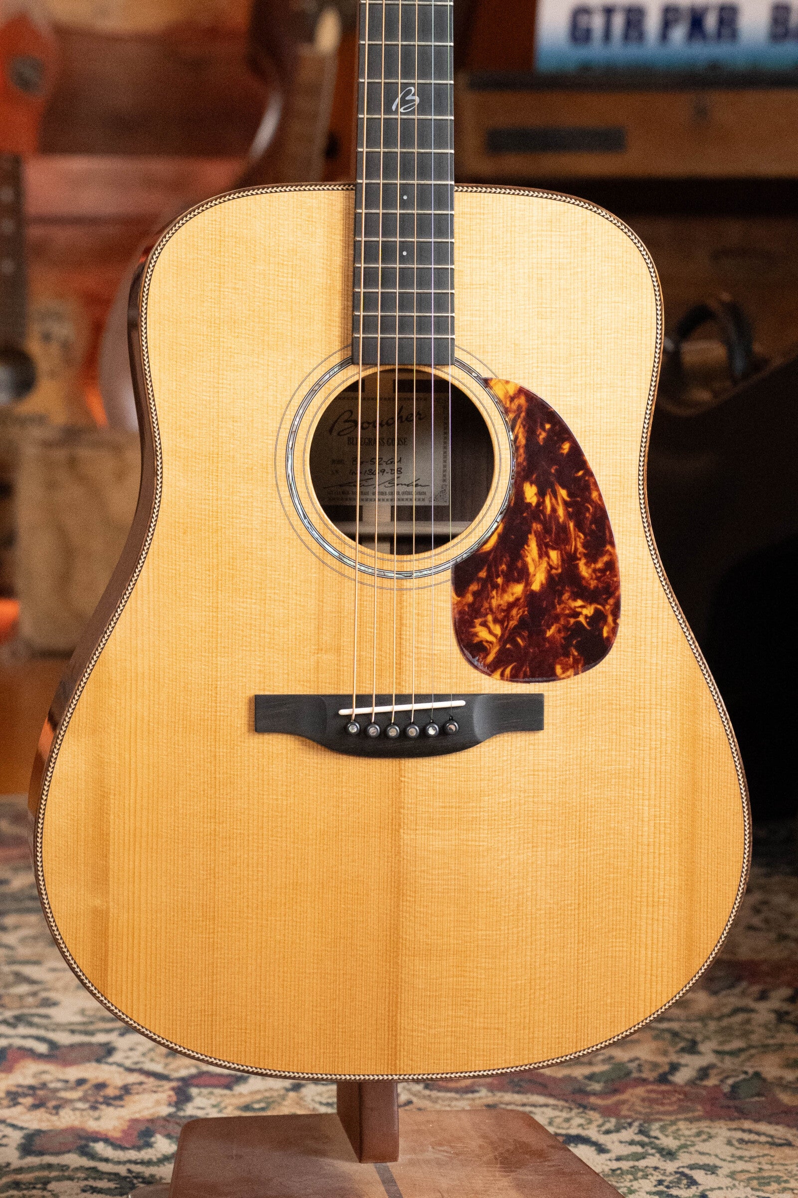 A close-up of the Boucher Guitars BG-52-GM Bluegrass Goose Series dreadnought, featuring a torrefied master grade Adirondack red spruce top and tortoiseshell pickguard, shown on a patterned rug with wooden background elements.