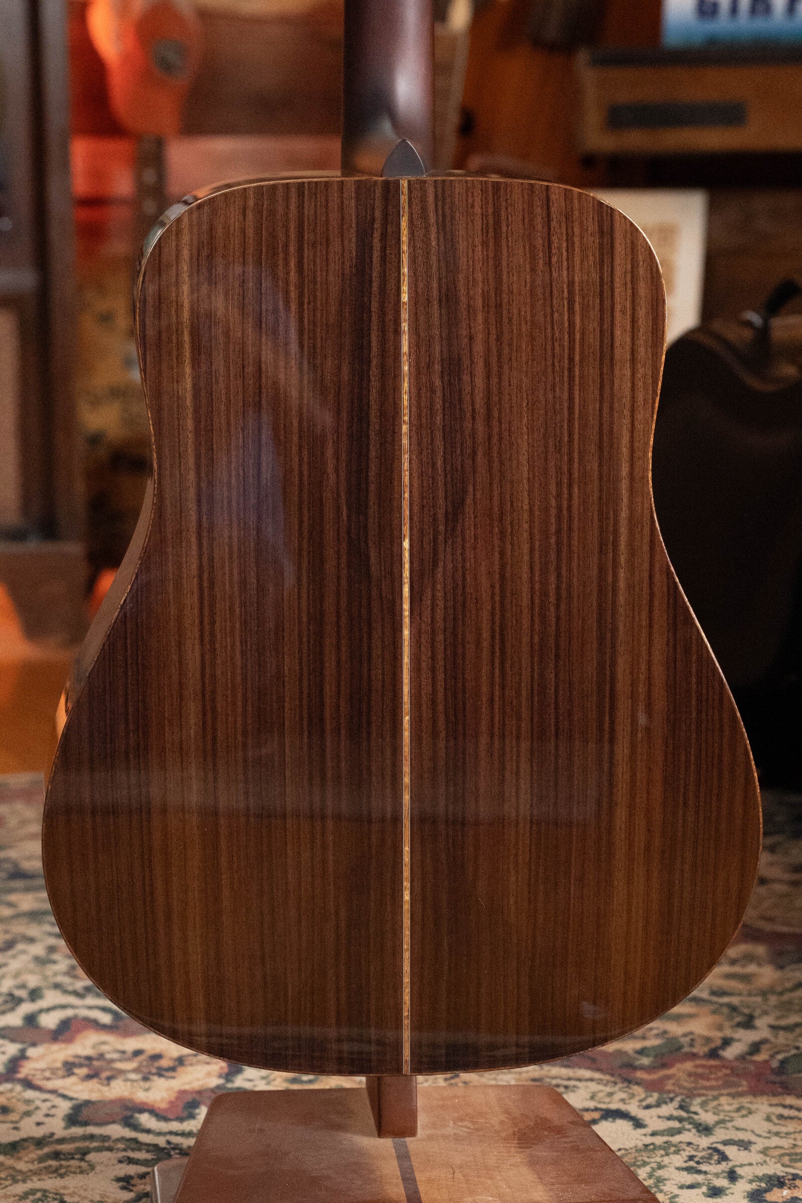 A close-up of the glossy East Indian rosewood back of the Boucher Guitars BG-52-GM Bluegrass Goose Dreadnought Acoustic Guitar, featuring a center strip. Other guitars and a patterned rug appear blurred in the background.