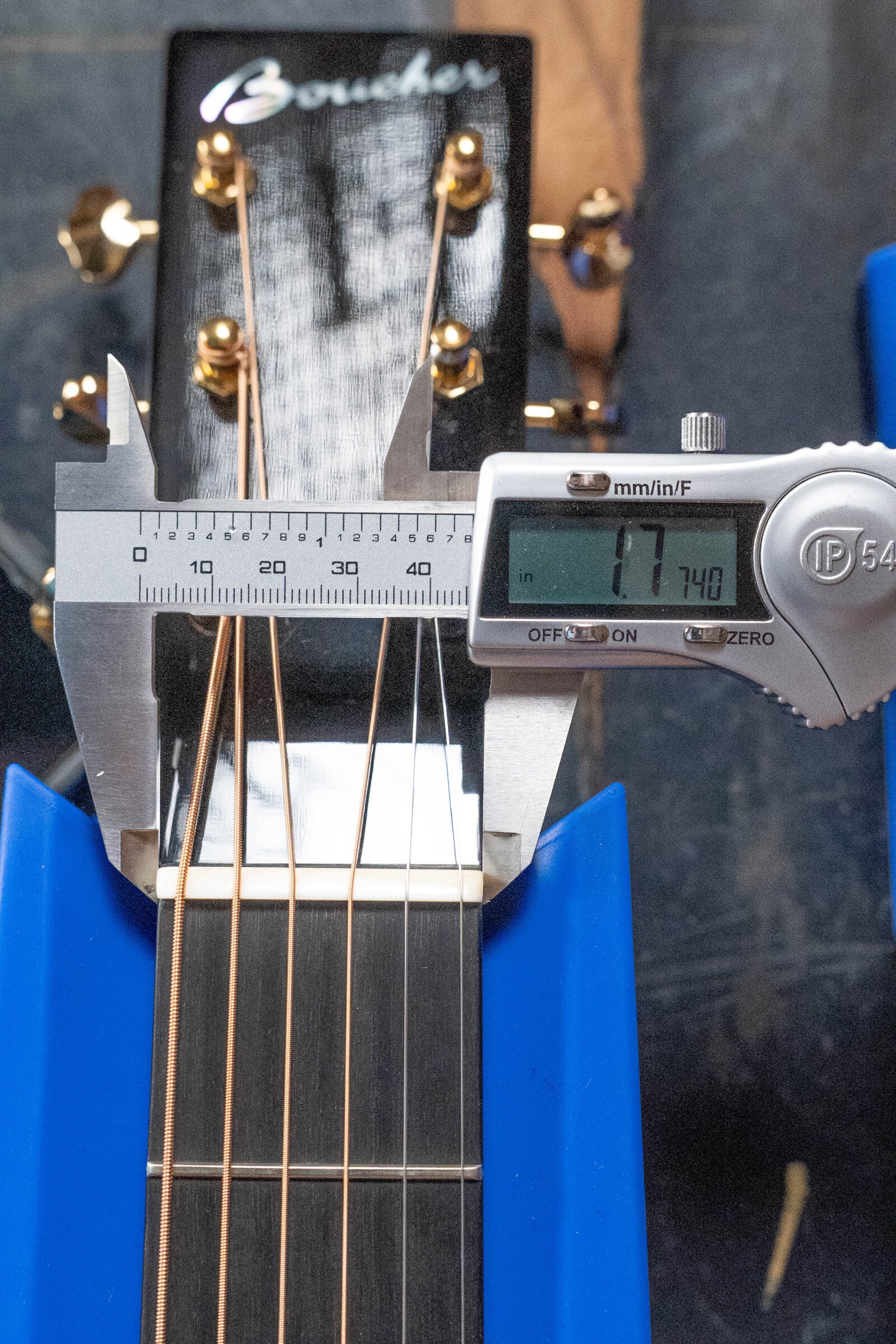 A digital caliper measures the nut width of the Boucher Guitars BG-52-GM Bluegrass Goose Dreadnought at 47.40 mm, with gold tuners and partial logo highlighting its East Indian rosewood and torrefied Adirondack spruce craftsmanship.