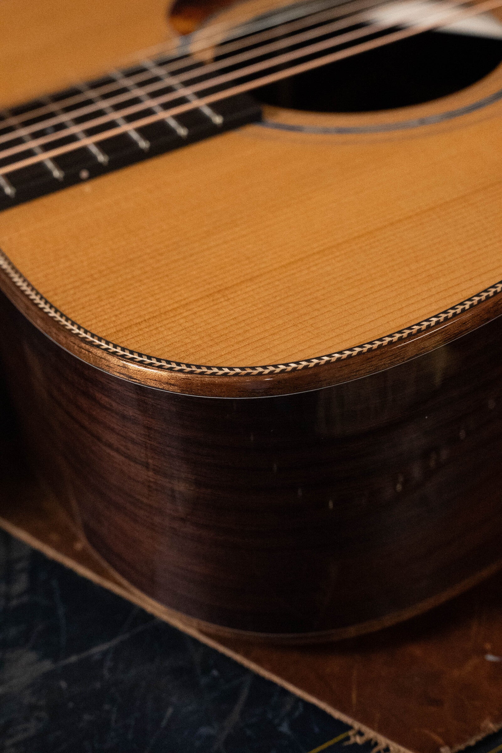 Close-up of the Boucher BG-52-GM Bluegrass Goose dreadnought by Boucher Guitars, showing East Indian rosewood grain, refined edge binding, part of the sound hole, strings, and fretboard.