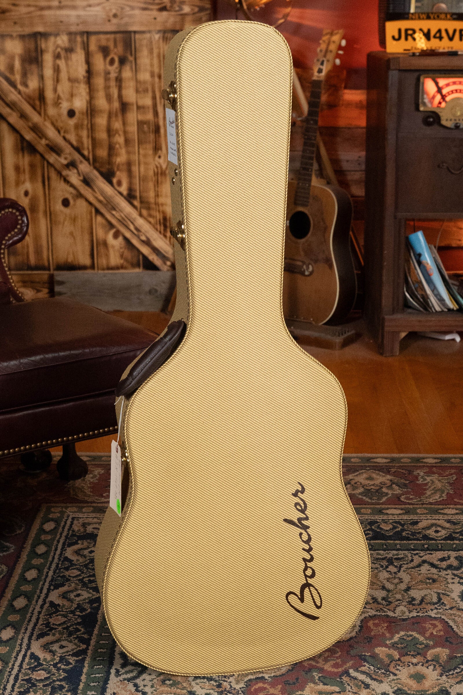 A Boucher BG-52-GM Bluegrass Goose acoustic guitar and a tan case marked Boucher stand upright on a patterned rug in a cozy wood-paneled room with vintage decor and a leather chair.