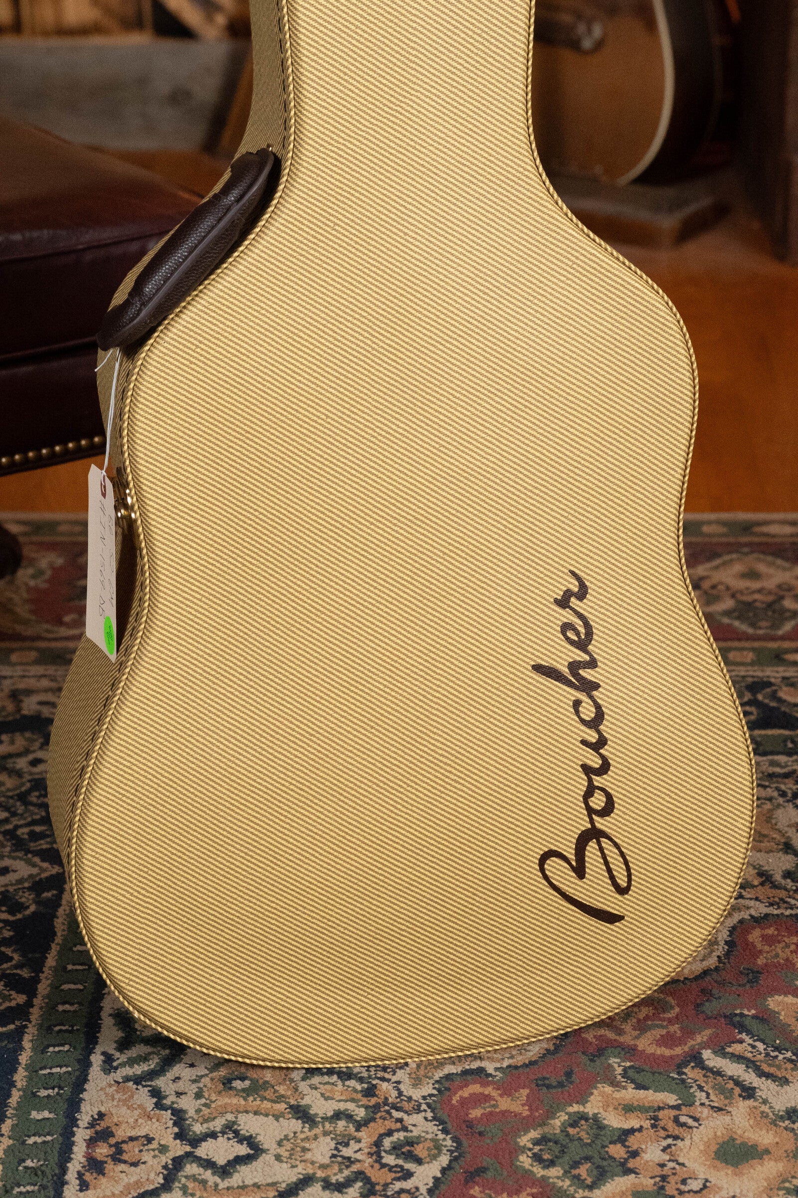 A Boucher Guitars beige textured case with black script sits on a patterned rug near a brown leather chair, next to a BG-52-GM Torrefied Master Grade Adirondack Red Spruce/East Indian Rosewood Bluegrass Goose Dreadnought.