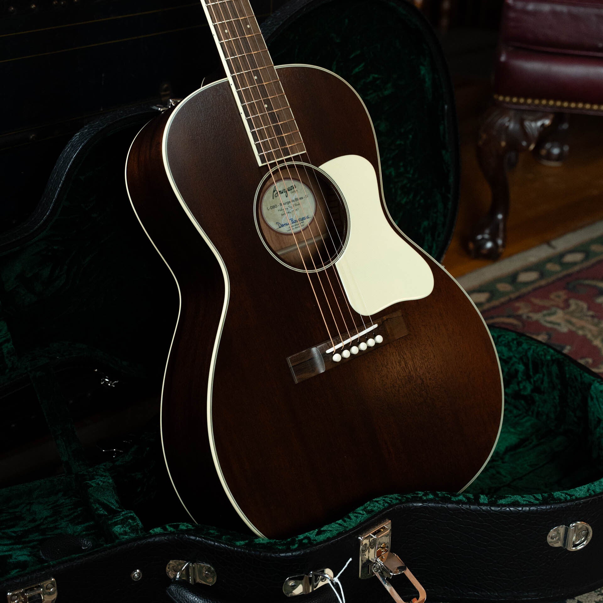 A Bourgeois Blues PSBB-LDBO-S Satin Professional Series All Mahogany OO Acoustic Guitar (#1197) with a white pickguard rests in an open green velvet-lined case on a wooden floor beside a patterned rug and furniture.