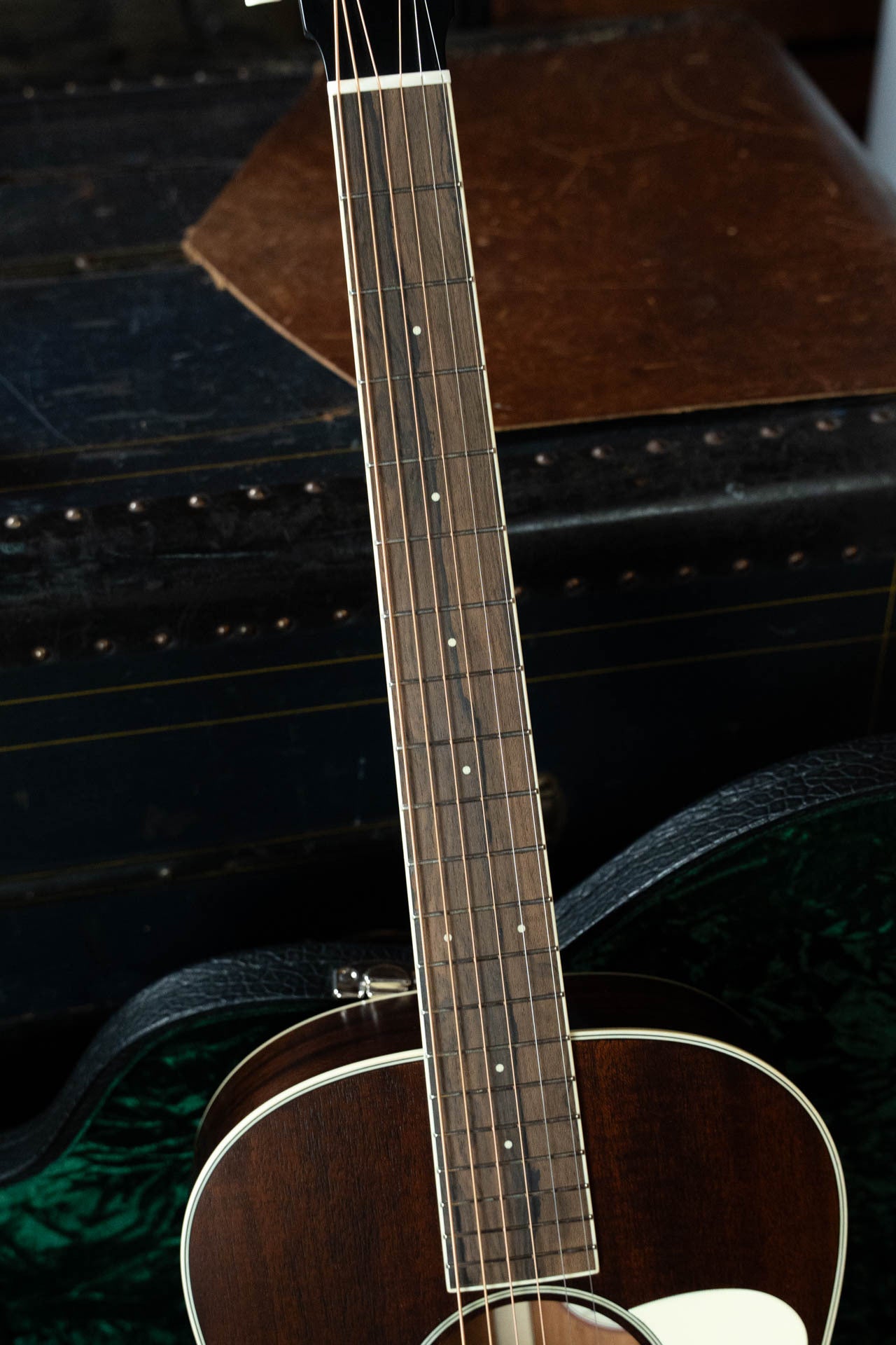 A close-up of the Bourgeois Blues PSBB-LDBO-S Satin Professional Series All Mahogany OO Acoustic Guitar #1197’s neck, fretboard, and strings, with a vintage trunk in the background.