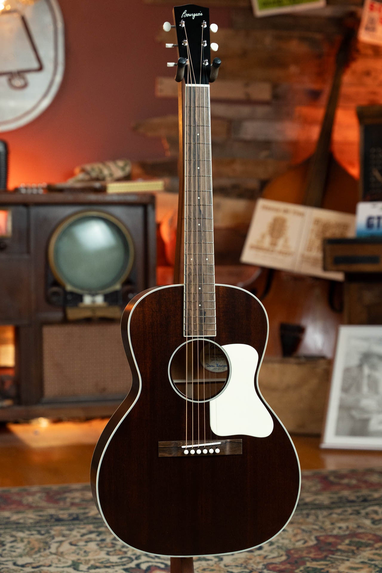 A Bourgeois Blues PSBB-LDBO-S Satin Professional Series All Mahogany OO Acoustic Guitar #1197 stands upright in a cozy room with vintage decor, featuring an old TV, radio, wooden walls, and a white pickguard.