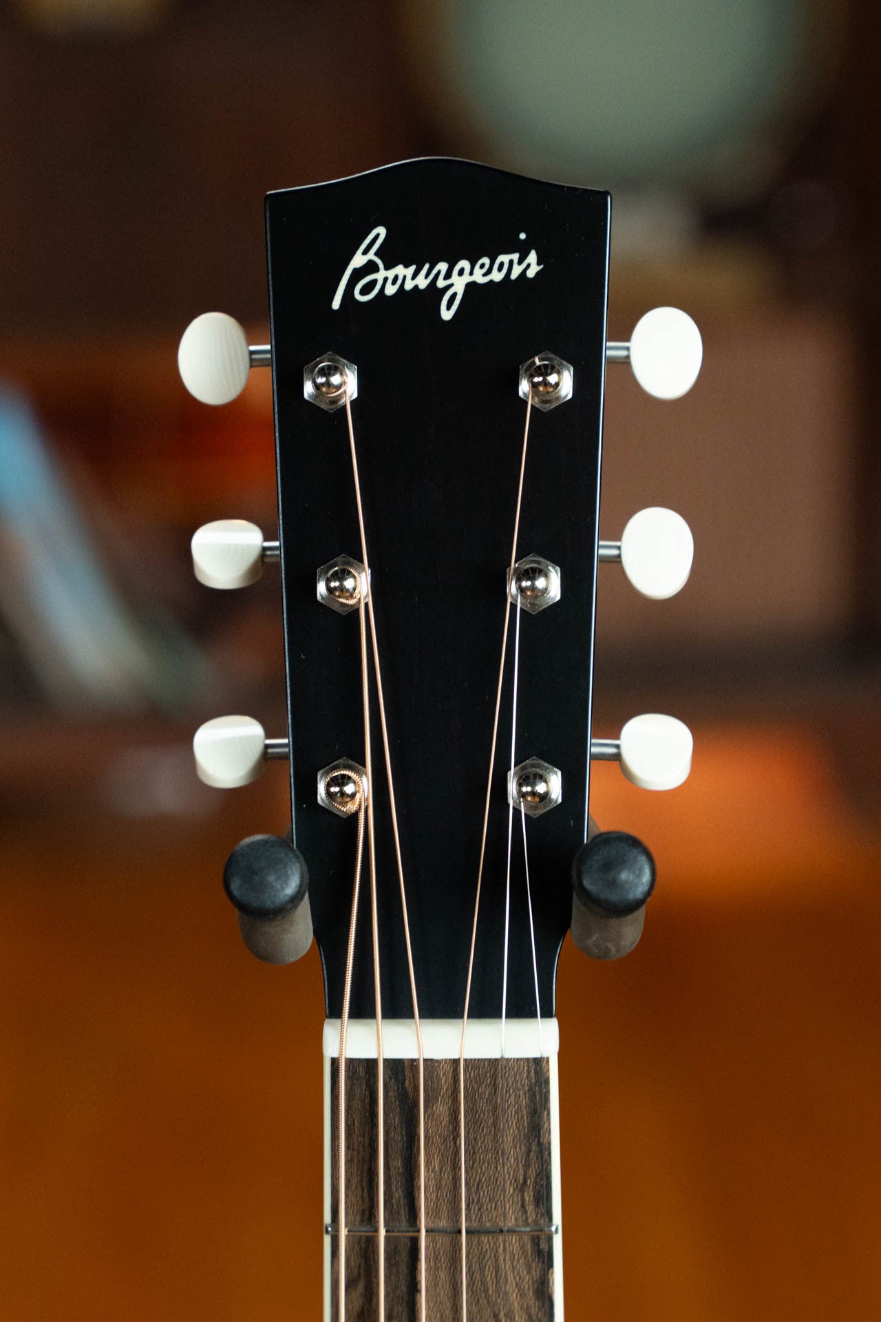 Close-up of a Bourgeois Blues PSBB-LDBO-S Satin Professional Series All Mahogany OO Acoustic Guitar #1197 headstock, highlighting six tuning pegs and strings, set against a softly blurred, warm brown background.