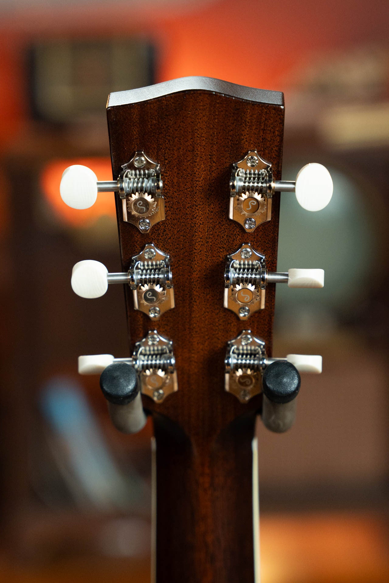 Close-up of the back of a Bourgeois Blues PSBB-LDBO-S Satin Professional Series All Mahogany OO Acoustic Guitar #1197 headstock, showing six chrome tuning pegs with white knobs against a warmly lit, blurred background.
