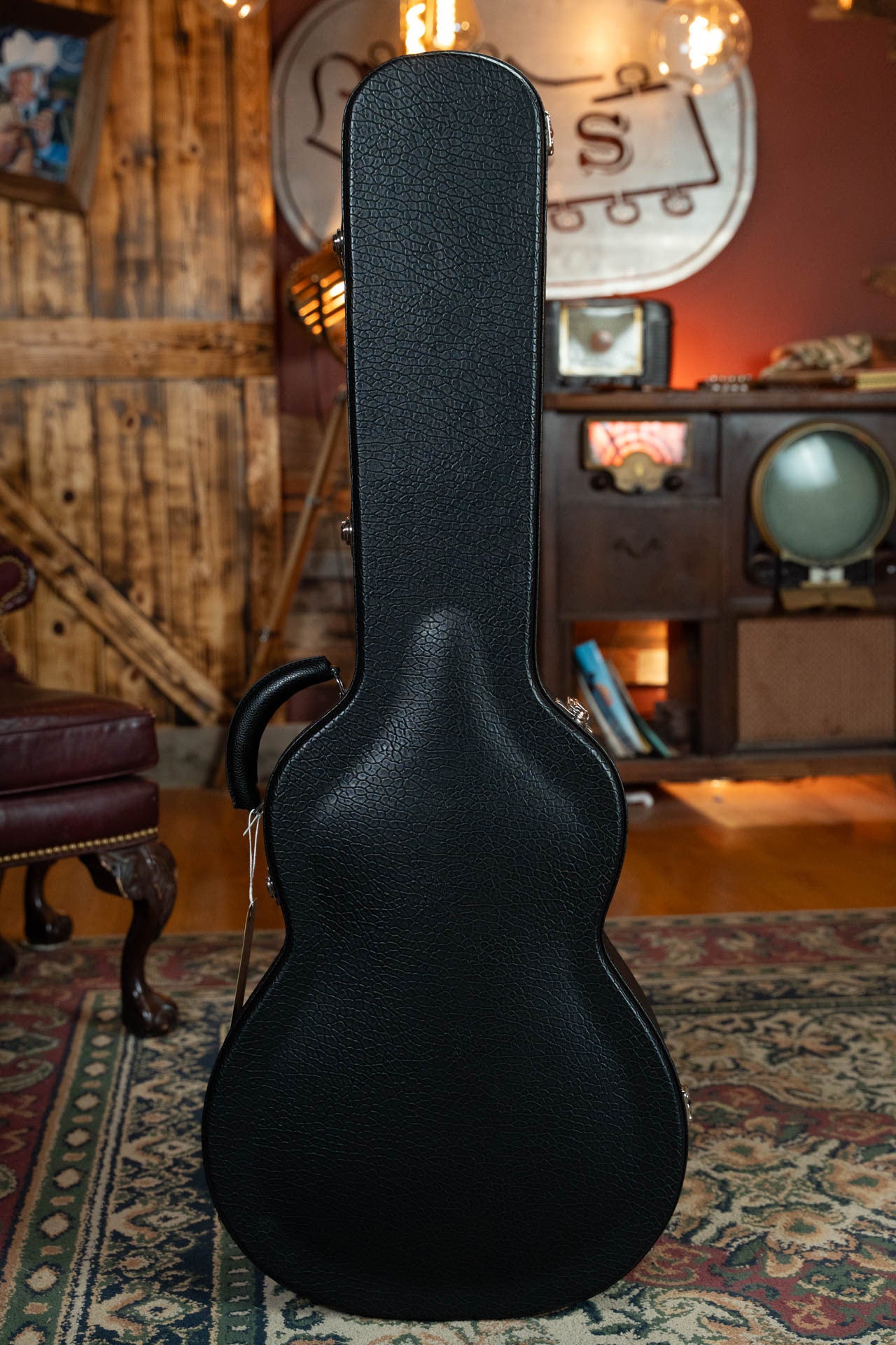 A black hard-shell guitar case stands upright on a patterned rug in a cozy room with vintage decor—the perfect fit for the Bourgeois Blues PSBB-LDBO-S Satin Professional Series All Mahogany OO Acoustic Guitar #1197.