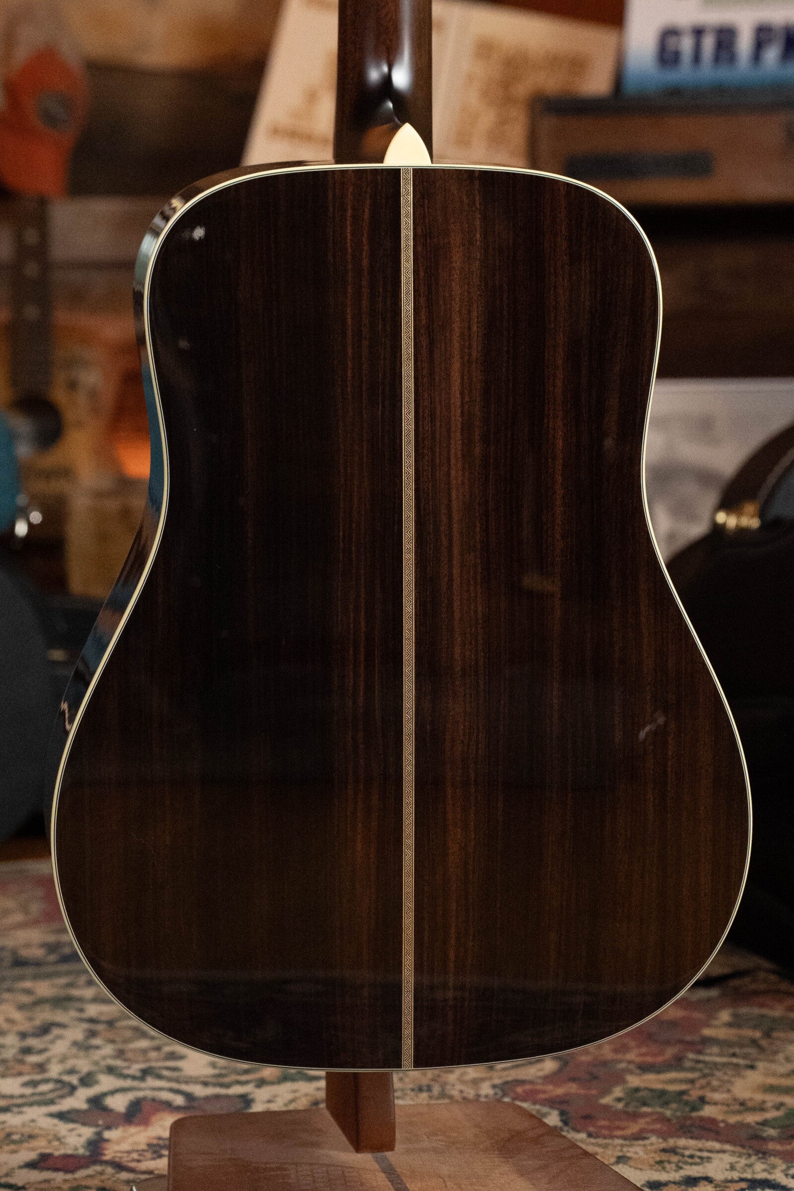 A close-up shows the back of a Bourgeois D Vintage/TS Touchstone Series Dreadnought Acoustic Guitar #7014 with a glossy dark wood finish and light center strip, resting on a stand. Wooden shelves and guitar cases fill the background.