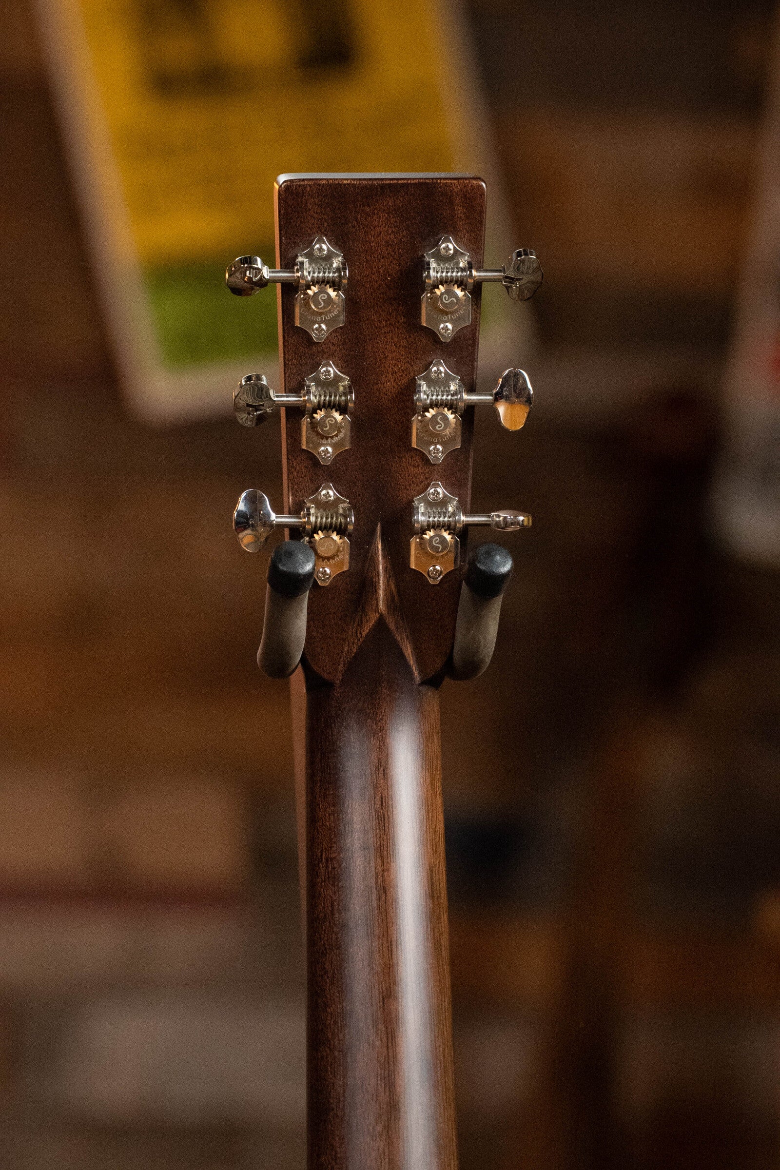 Close-up of the back of a Bourgeois D Vintage/TS Touchstone Series Dreadnought Acoustic Guitar #7014 headstock, displaying six tuning pegs and part of the guitar stand, with posters and a brick wall softly blurred in the background.