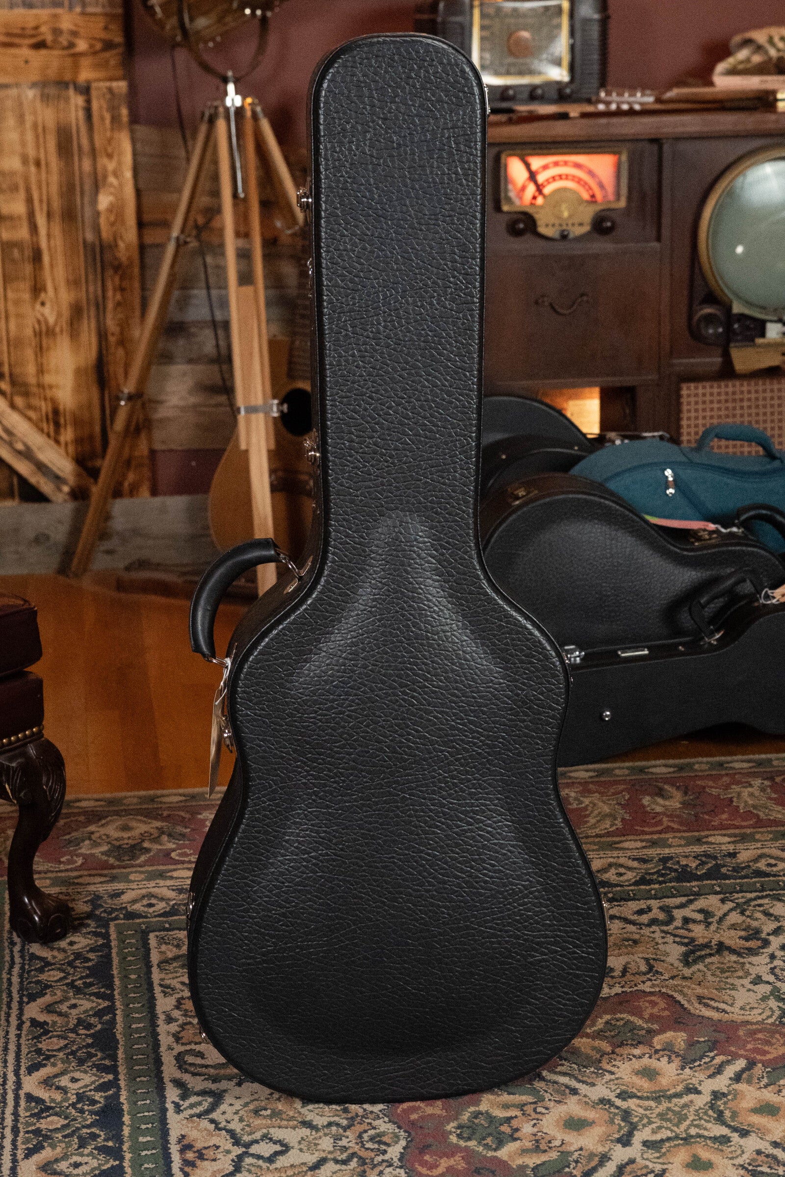 A black hard-shell guitar case stands upright on a rug, with vintage audio gear and a tripod behind—ideal for your Bourgeois D Vintage/TS Touchstone Series Dreadnought Acoustic Guitar #7014.