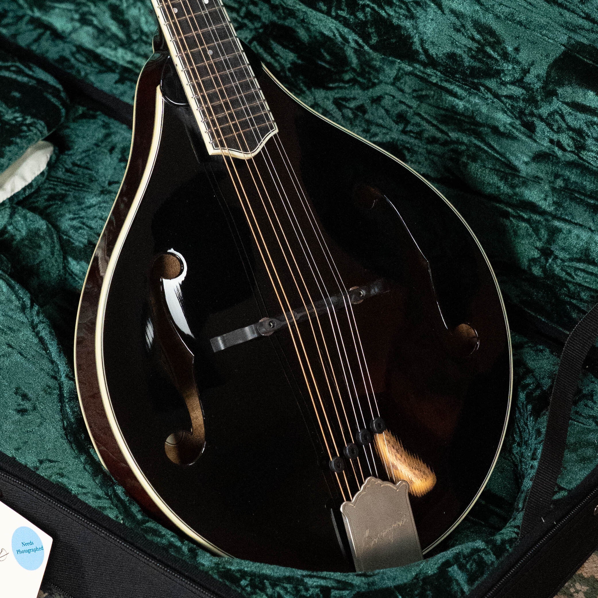 A Bourgeois M5-A-BT Aged-Tone Black Top A-Style Mandolin (#2020) with an Adirondack spruce top and light trim rests in a green velvet-lined case. The black instrument, viewed from above, has f-holes and multiple strings.