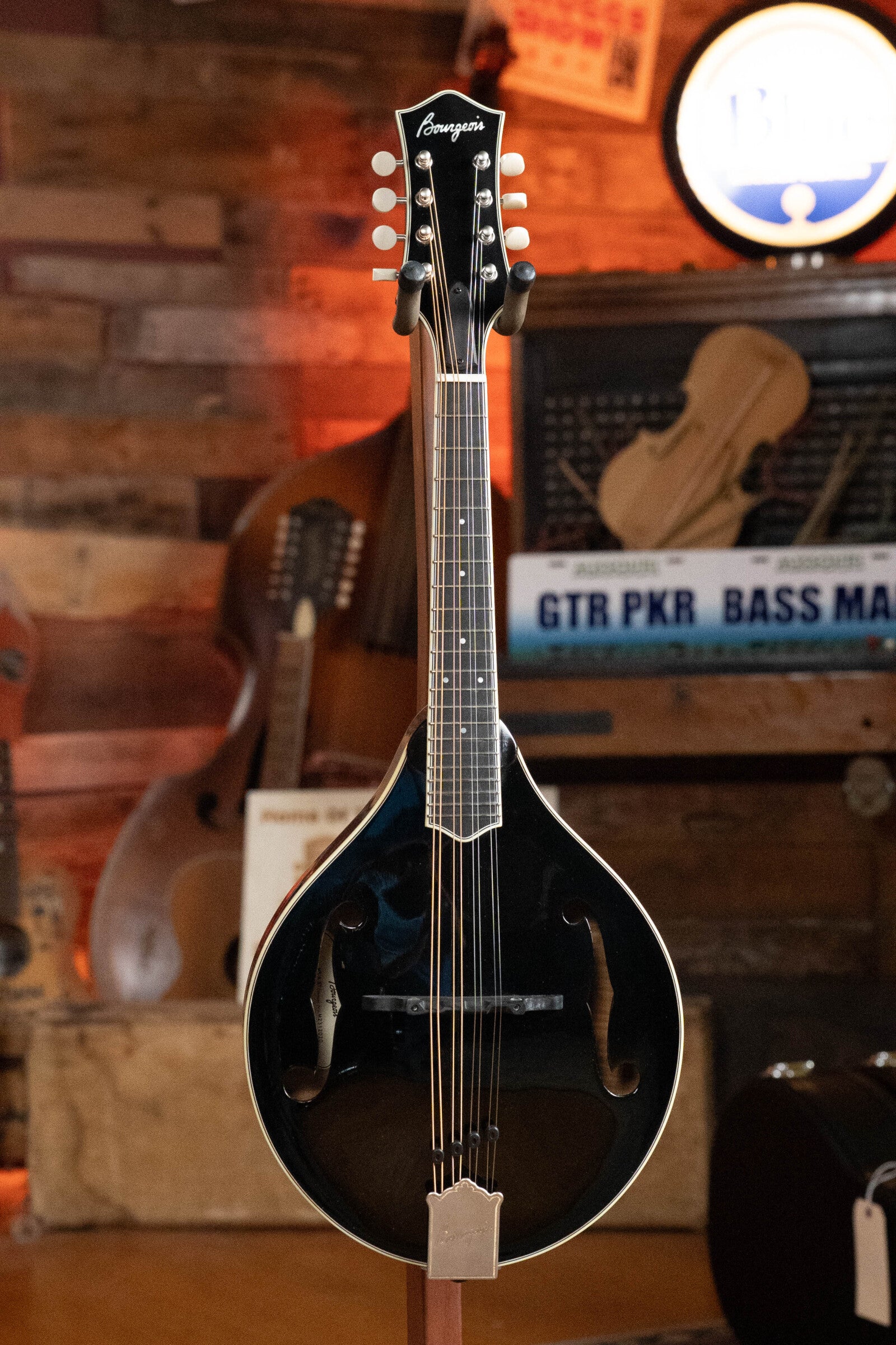 The Bourgeois M5-A-BT Aged-Tone Black Top A-style Mandolin #2020 by Bourgeois stands upright on a display stand before a rustic wooden wall, with blurred instruments and signs in the background.