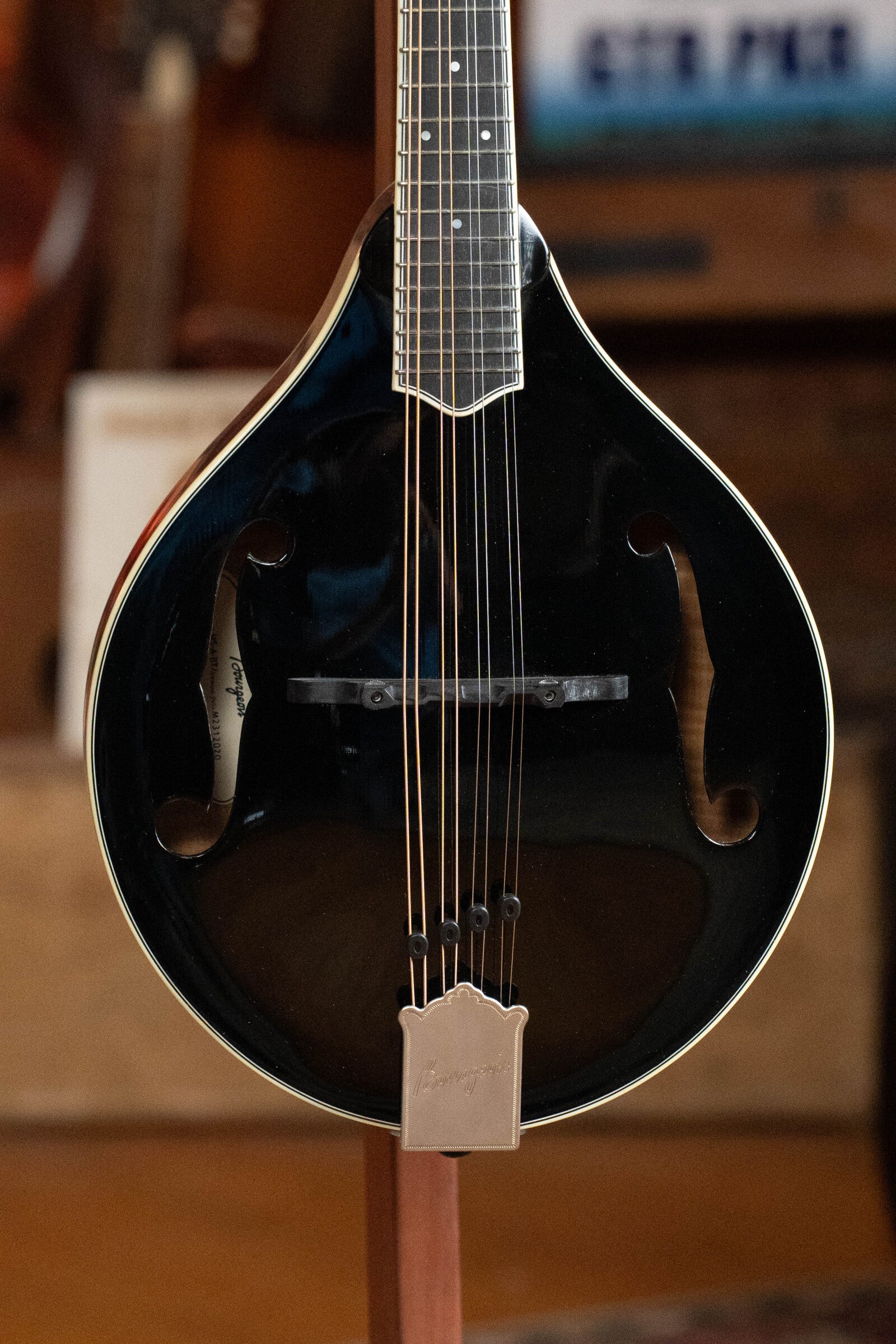 A close-up of the Bourgeois M5-A-BT Aged-Tone Black Top A-Style Mandolin #2020 on a stand, displaying its strings, f-holes, and glossy Adirondack spruce top by Bourgeois, with a blurred background.