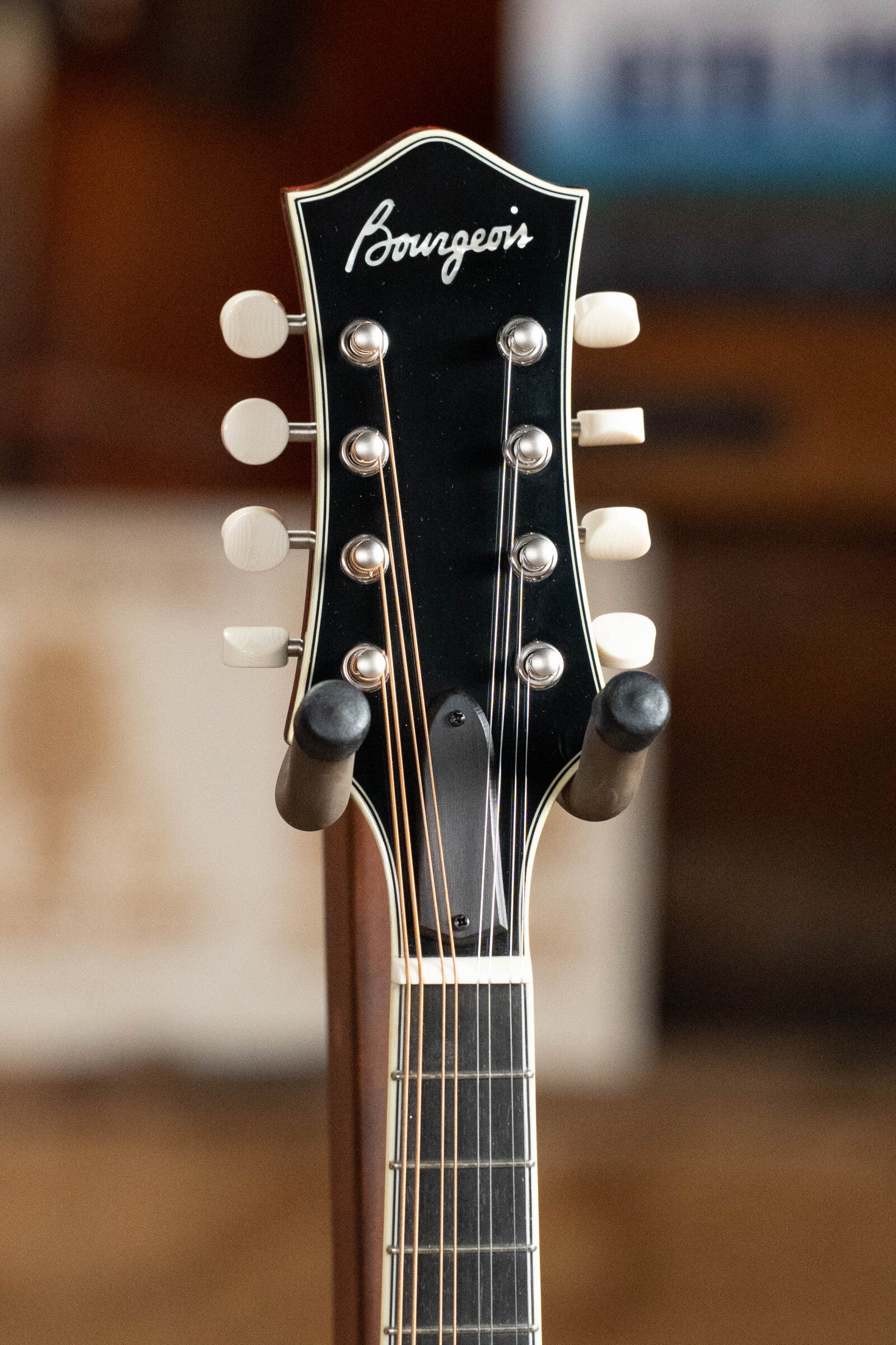 Close-up of the Bourgeois M5-A-BT Aged-Tone Black Top A-Style Mandolin #2020 headstock, featuring six tuning pegs with white buttons and the Bourgeois logo, resting on a stand with a softly blurred background.