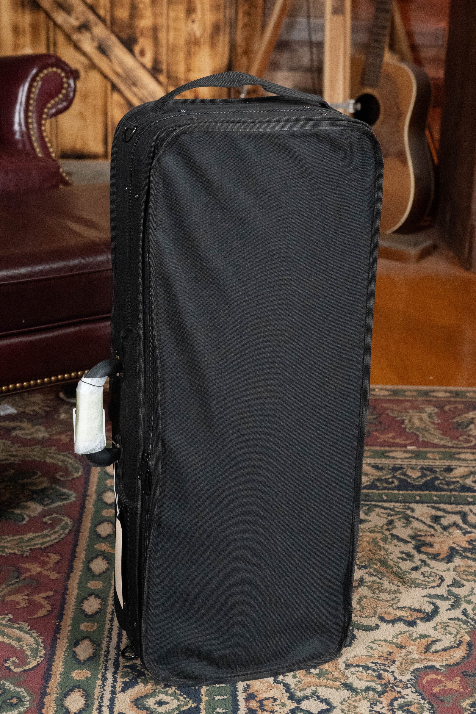 A black rectangular Bourgeois case stands upright on a patterned rug, ideal for safely storing your Bourgeois M5-A-BT Aged-Tone Black Top A-Style Mandolin #2020, with a leather chair and acoustic guitar in the background.
