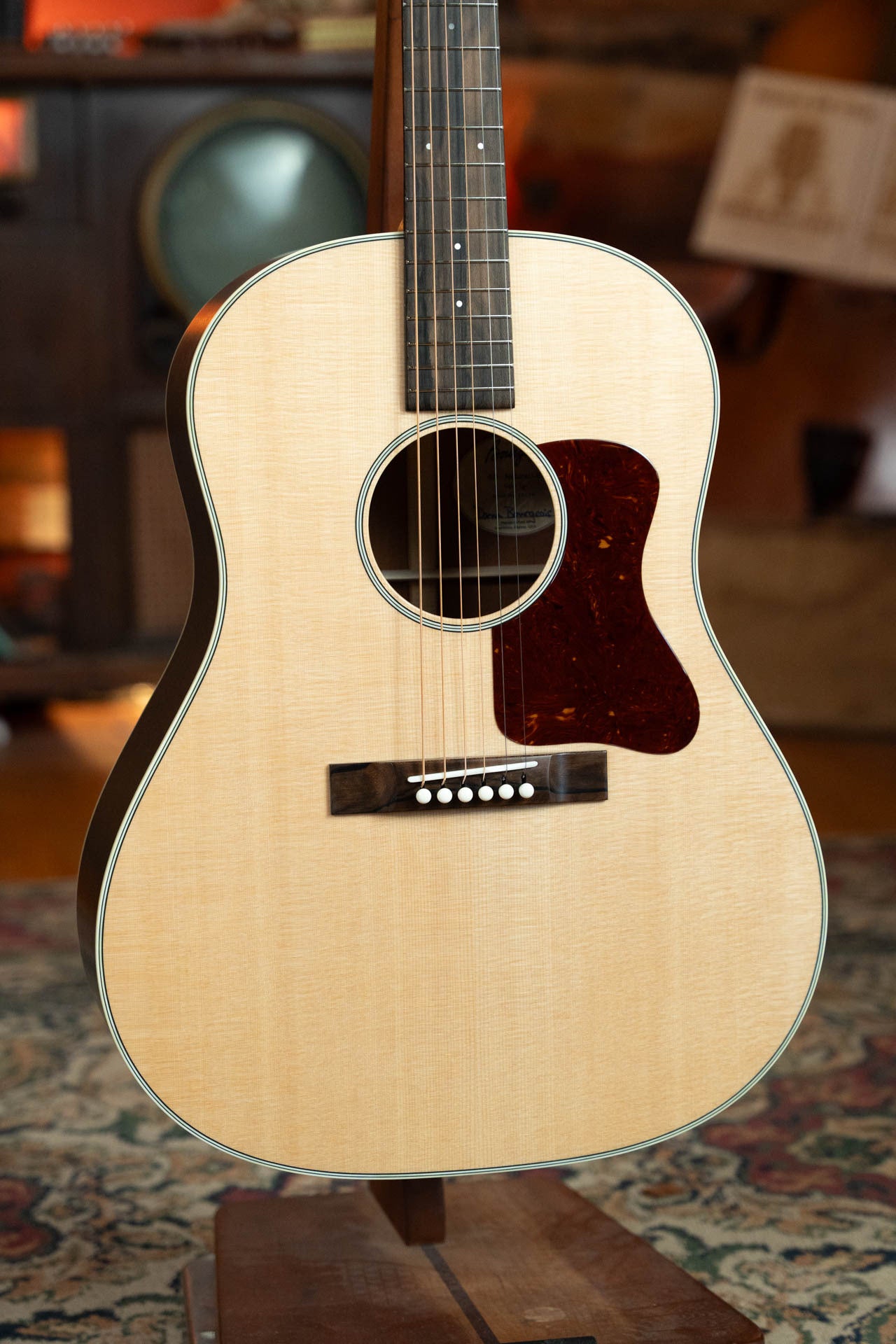 A close-up of the Boucher Guitars Bourgeois PSN-SD-S Satin Professional Series The Natural Sitka/Mahogany Slope Shoulder Dreadnought Acoustic Guitar #1174 on a stand indoors, with a light wood body and dark pickguard on a patterned rug.
