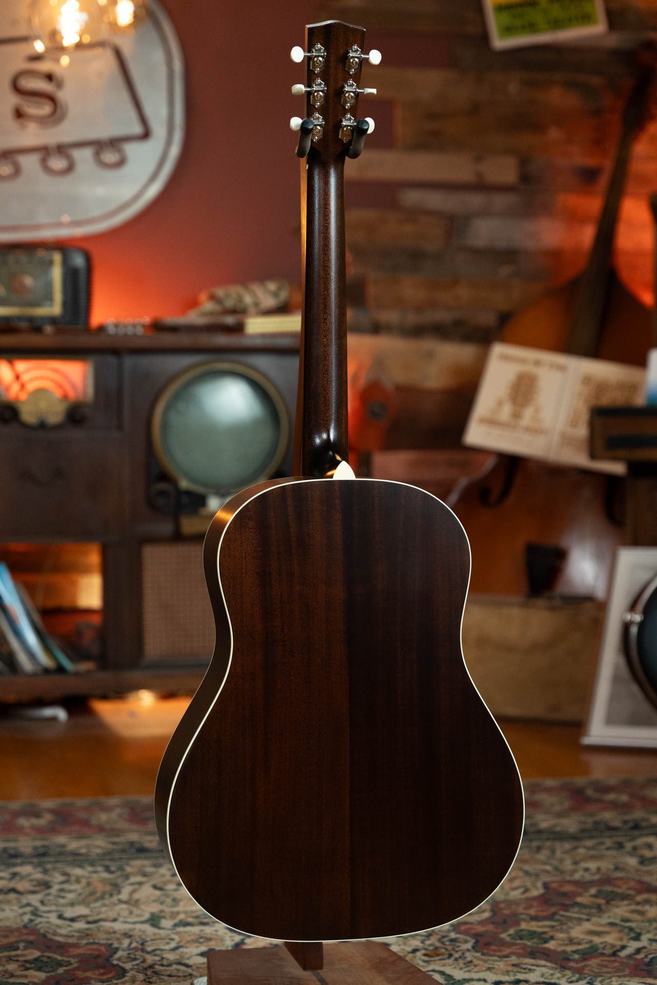 A Boucher Guitars PSN-SD-S Satin Professional Series Slope Shoulder Dreadnought #1174 with Sitka/Mahogany is displayed upright in a cozy, vintage-style room with a patterned rug, old radios, and various instruments.