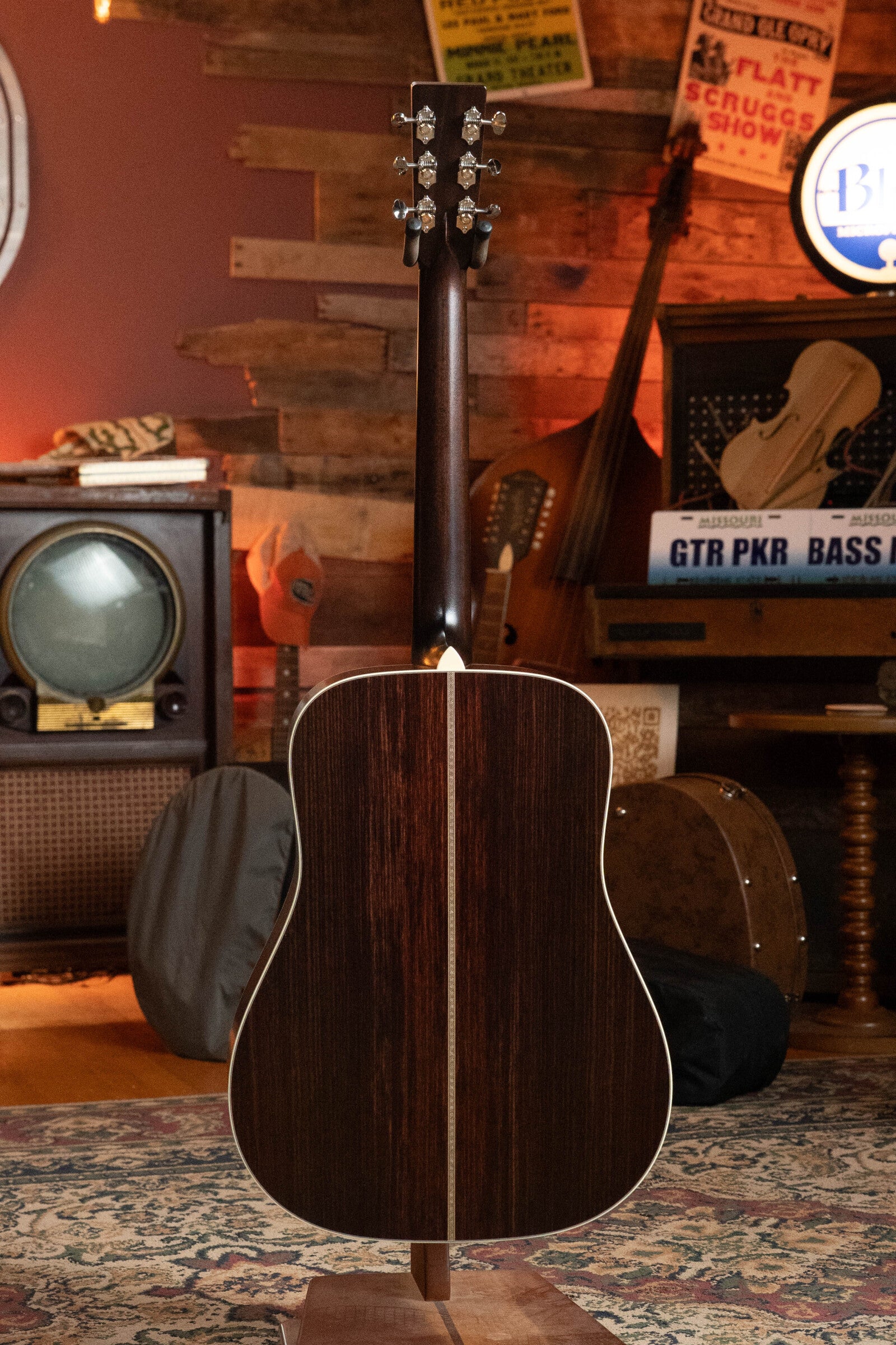 A Bourgeois PSV-D-S Professional Series Sitka/Rosewood Vintage Satin Dreadnought Acoustic Guitar #1124 stands on a stand in a cozy, vintage-style room with rustic decor, musical instruments, an old TV, and posters.