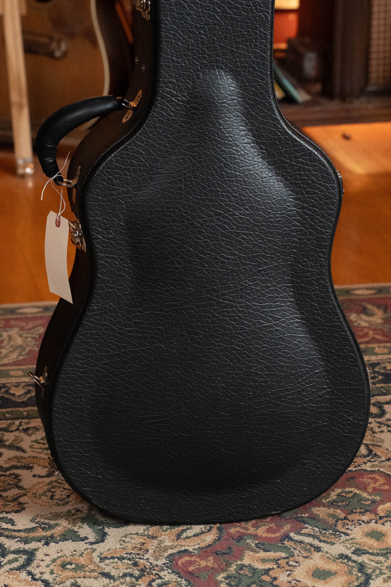 A black, textured guitar case with a handle and white tag stands upright on a patterned rug in a warmly lit room with wood floors, musical instruments, and a Bourgeois PSV-D-S Professional Series Sitka/Rosewood Dreadnought (#1124) in the background.