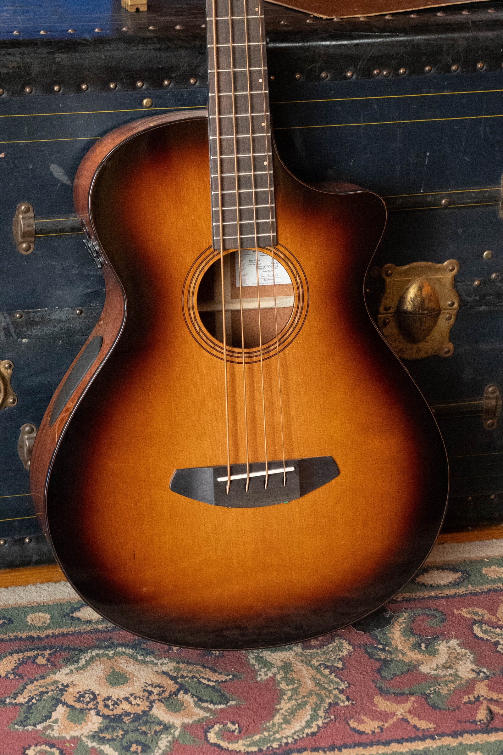 A Breedlove Organic Solo Pro Concerto Red Cedar/African Mahogany Edgeburst Cutaway Acoustic Bass w/ Fishman Pickup (#6054) by Breedlove leans against a vintage blue trunk with brass hardware, resting on a patterned rug. Part of its neck and body are visible.