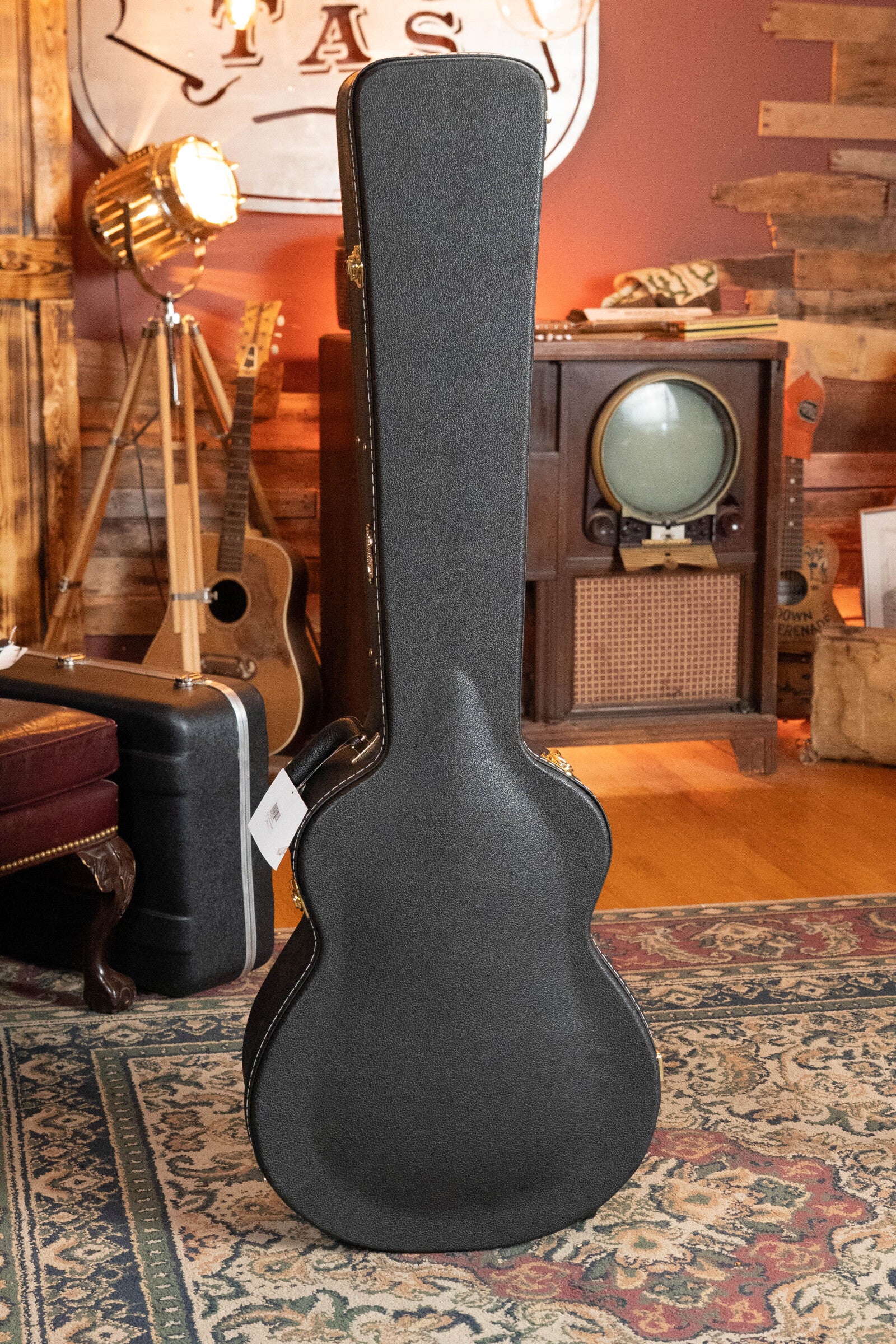 A Breedlove Organic Solo Pro Concerto acoustic bass rests upright in a cozy, vintage-themed room featuring a patterned rug, old TV, suitcase, retro tripod lamp, and black guitar case.