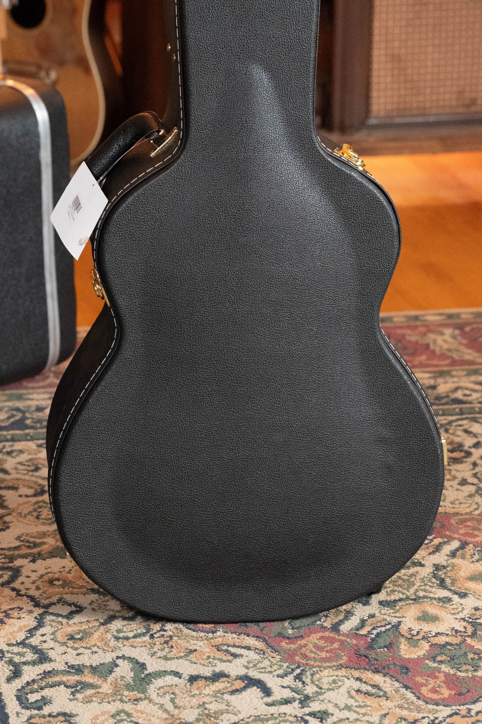A black hard guitar case with a white tag stands on a patterned rug, with wooden flooring and parts of a Breedlove Organic Solo Pro Concerto Red Cedar/African Mahogany Edgeburst Cutaway Acoustic Bass visible in the background.