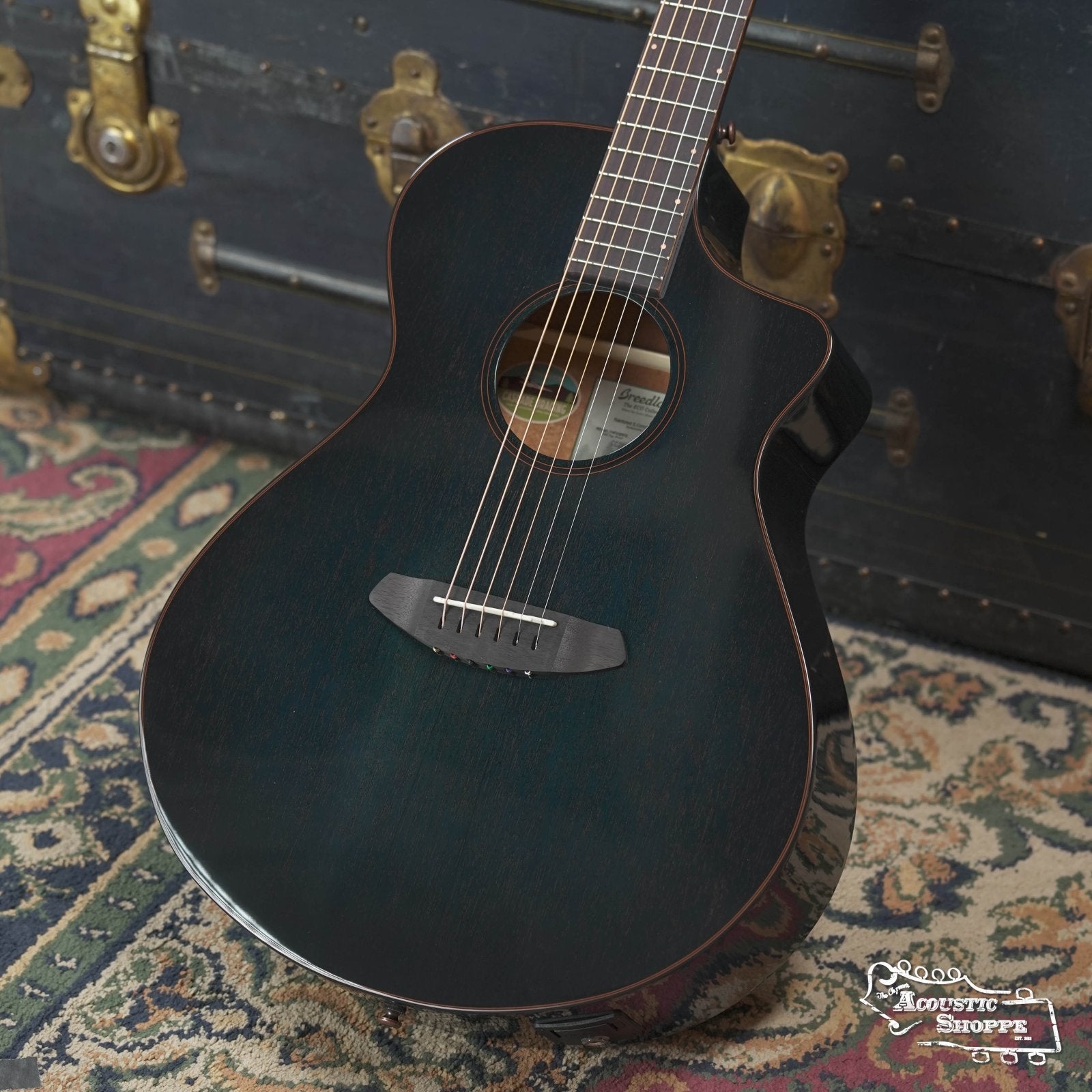 A stylish Breedlove Rainforest S Concert Papillon All Mahogany Cutaway Acoustic Guitar w/Fishman Pickup sits on a patterned rug before a vintage trunk, with The Acoustic Shoppe logo at the lower right corner of the image.